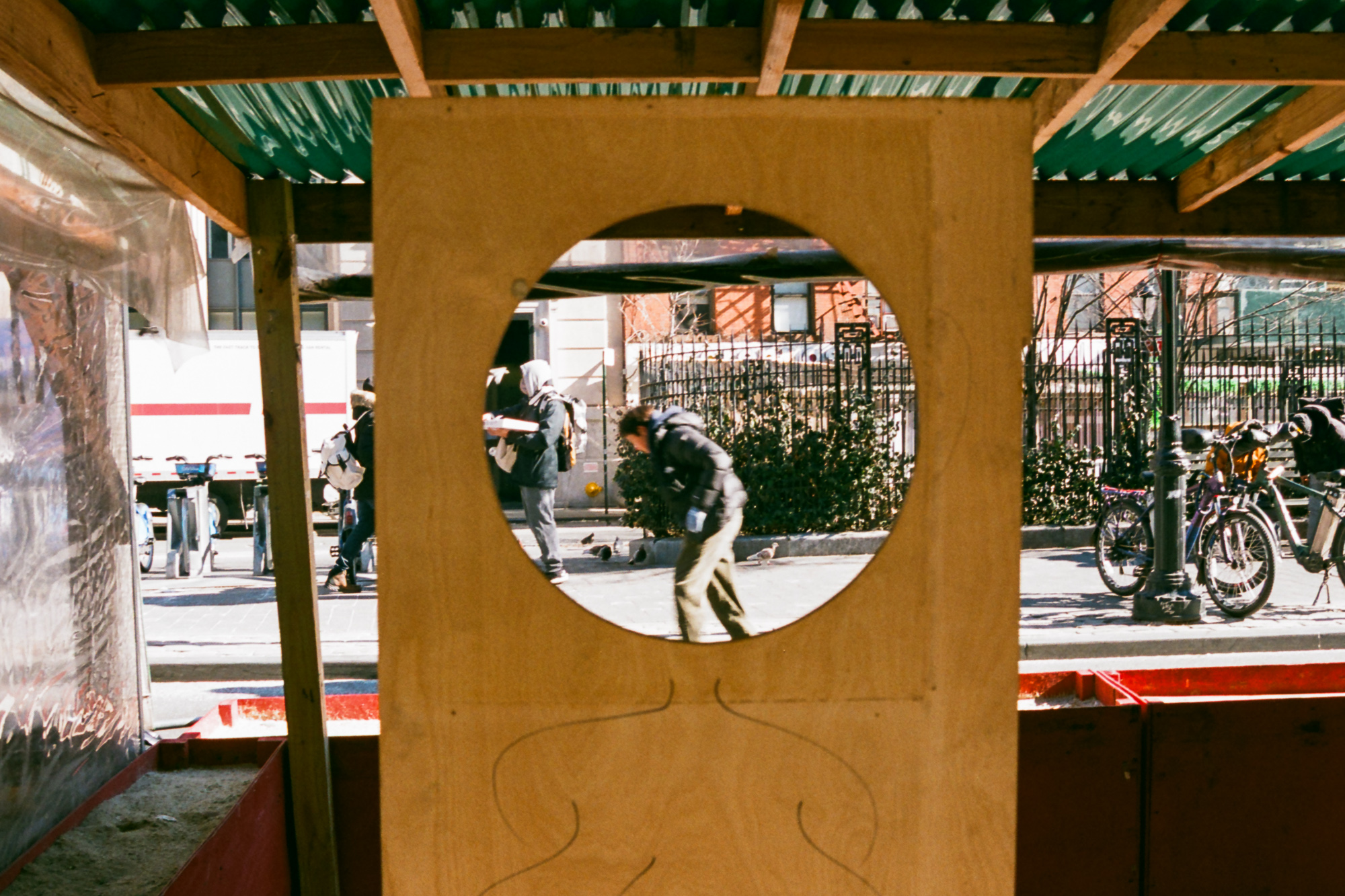 A plywood barrier under a green-roofed construction scaffold has a large circular hole cut into it, framing a view of the sidewalk beyond where a man in a black puffer jacket walks bent forward and a hooded pedestrian carries a white box. Bicycles, an iron park fence and a white box truck fill the scene on either side of the wooden panel.