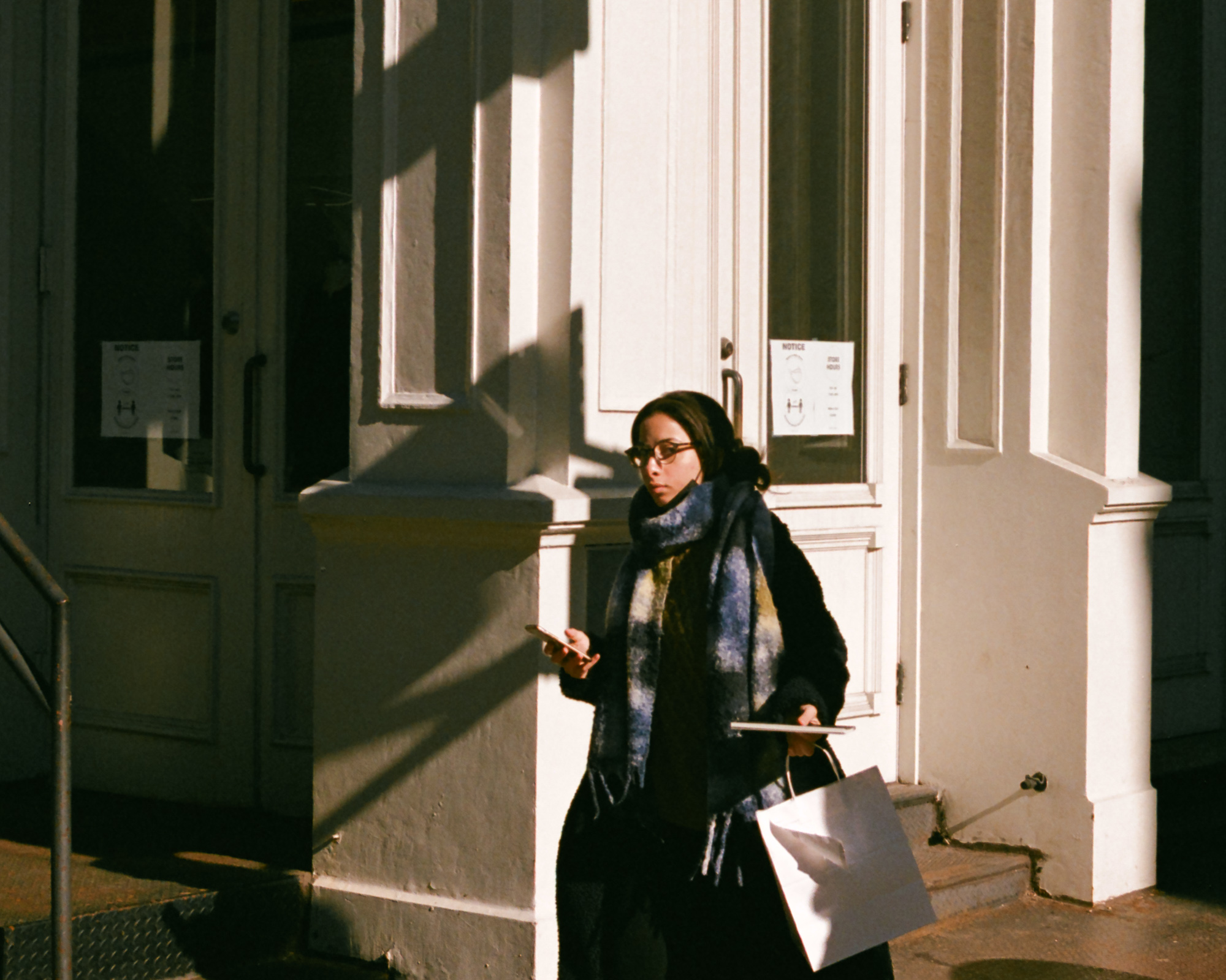 A woman in glasses, a black coat and a thick blue and yellow plaid scarf walks past a cream-painted storefront in hard morning sunlight, holding a phone in one hand and a white shopping bag and slim case in the other, turning her head toward the camera. Diagonal shadows from the building's cornices cut across the wall behind her, and a NOTICE and store hours sign is taped inside the glass door.