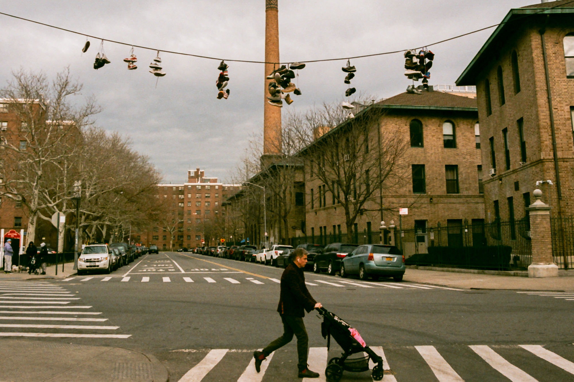 A man in a dark jacket pushes an empty stroller across a wide crosswalk on an overcast day, while dozens of pairs of sneakers hang by their laces from a power line strung across the intersection above him. Brick apartment buildings and a tall industrial smokestack rise behind parked cars lining both sides of the street.