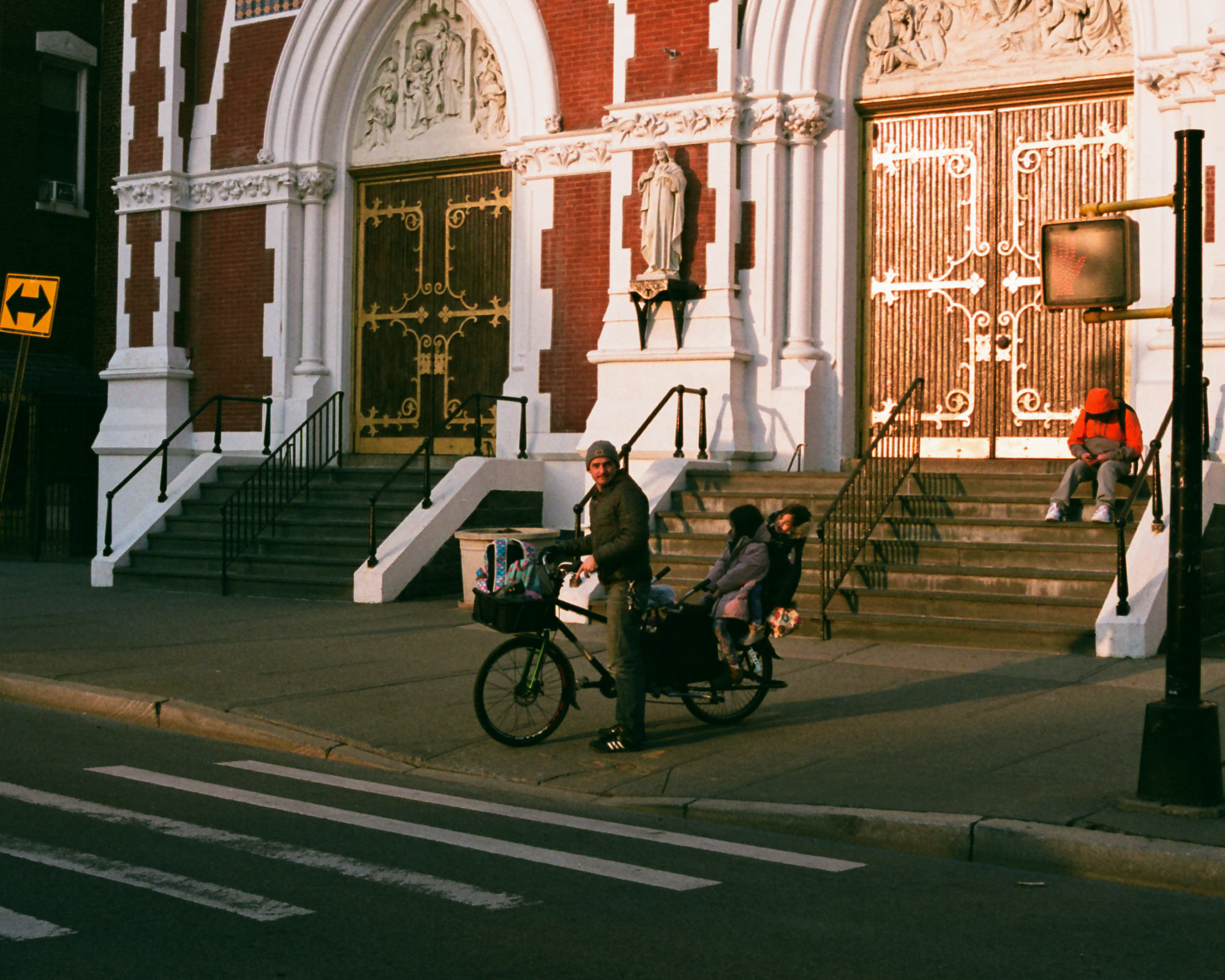 A man in a knit cap and dark jacket stands astride a long cargo bike on a sidewalk in front of a brick church with white stone arches and gold-painted doors, two bundled children seated behind him, while a person in an orange hooded jacket sits alone on the church steps to the right.