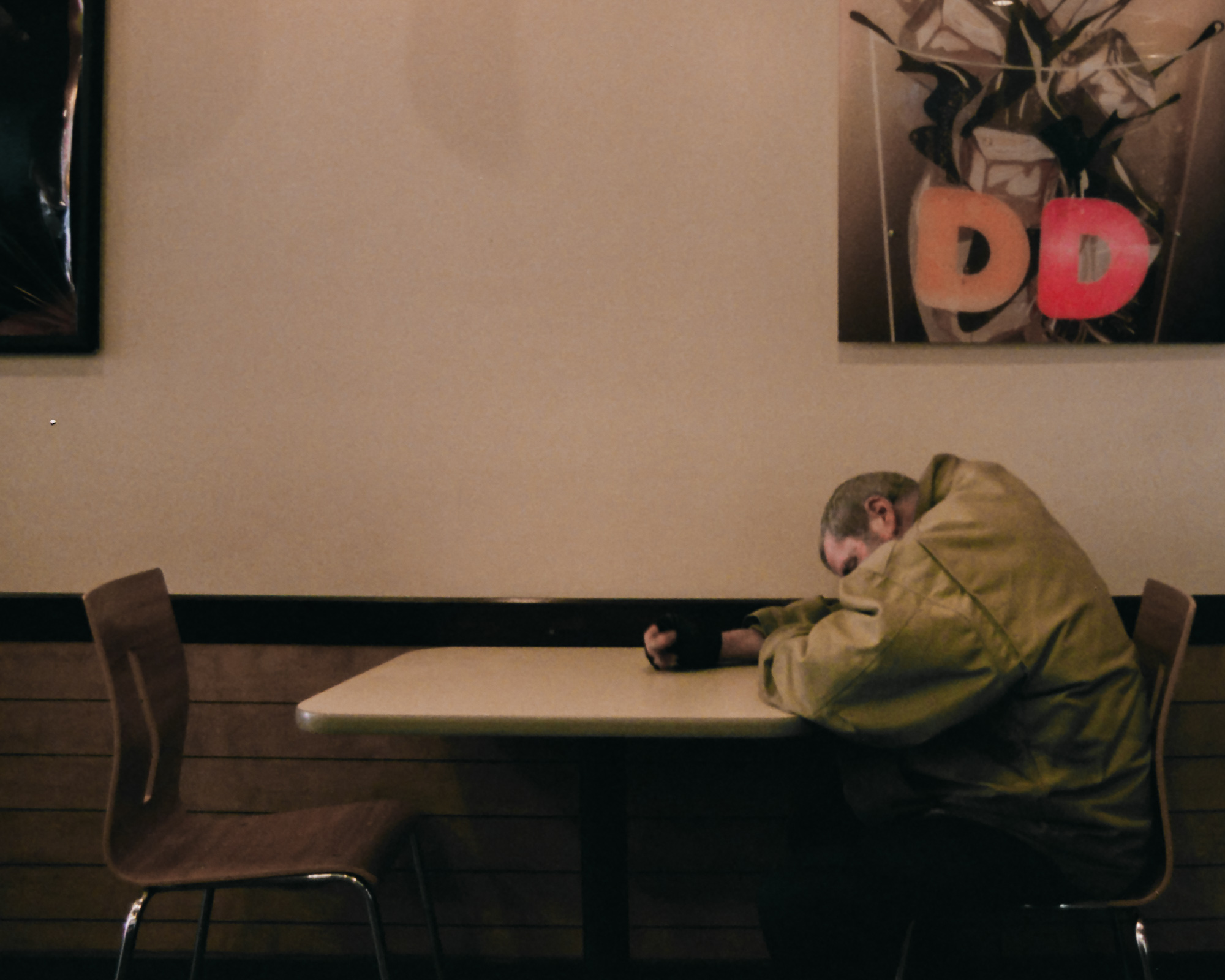 A man in a bulky olive-green coat rests his head face down on his folded arms at a laminate table inside a donut shop, wearing a black fingerless glove. An empty brown chair sits across from him, and framed coffee-themed artwork hangs on the beige wall above.
