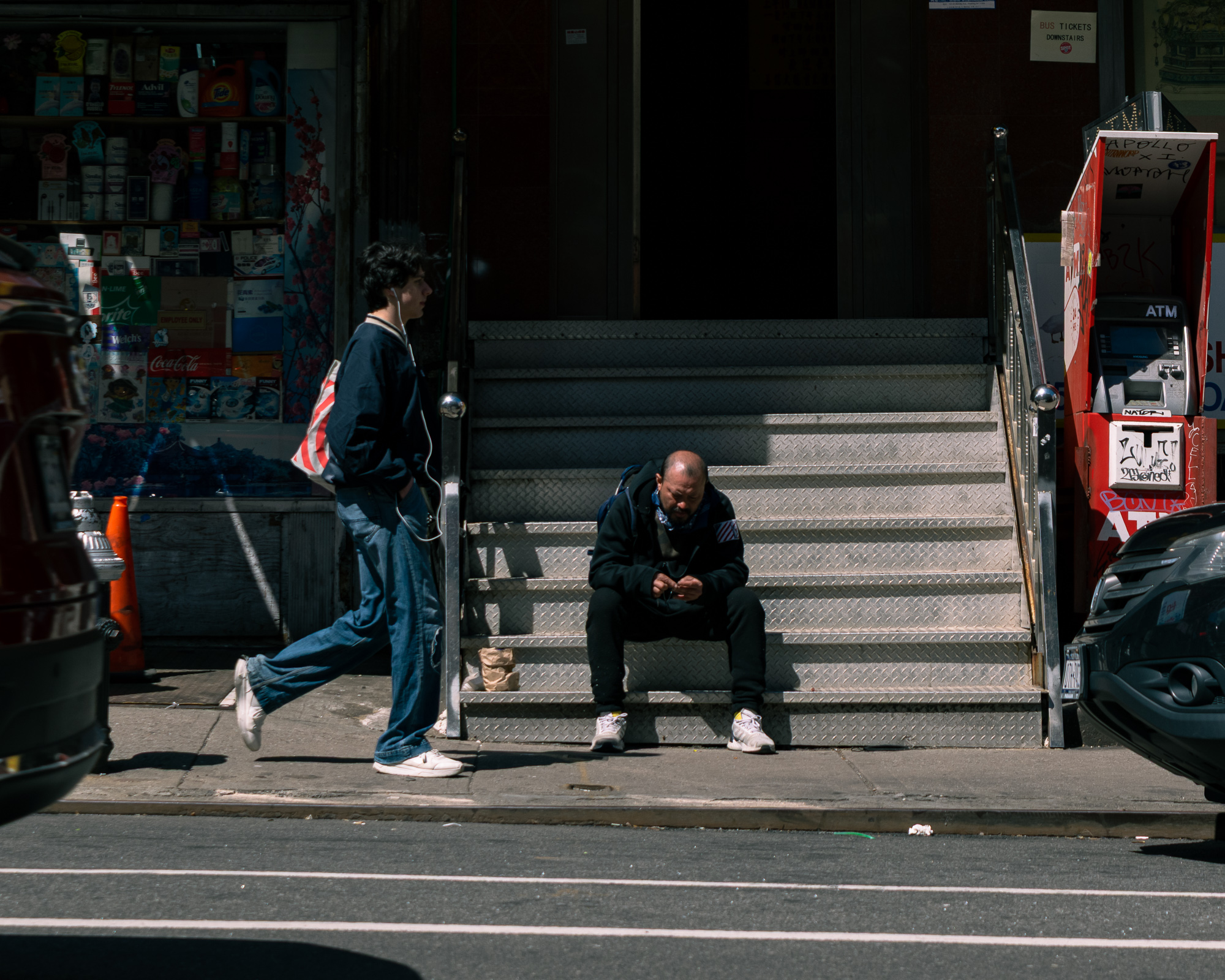 A man in a black hooded jacket sits hunched forward on metal diamond-plate subway steps in bright midday sun, while a young man in a navy jacket and wide jeans with earbuds walks past on the sidewalk in front of a bodega window stocked with detergent and soda signs. A graffiti-covered red ATM booth stands at the right of the stairwell.