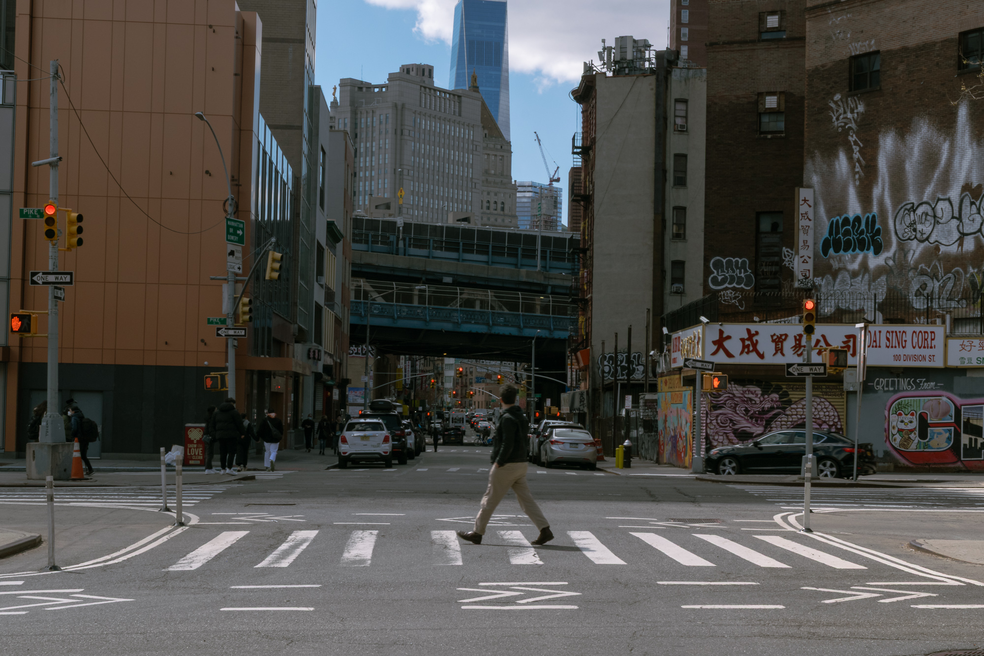 A man in a dark jacket and tan trousers walks alone across a wide painted crosswalk at a Chinatown intersection, with a red traffic signal overhead, the blue steel Manhattan Bridge approach spanning the street behind him, and the Dai Sing Corp sign and graffiti-covered brick wall to the right.