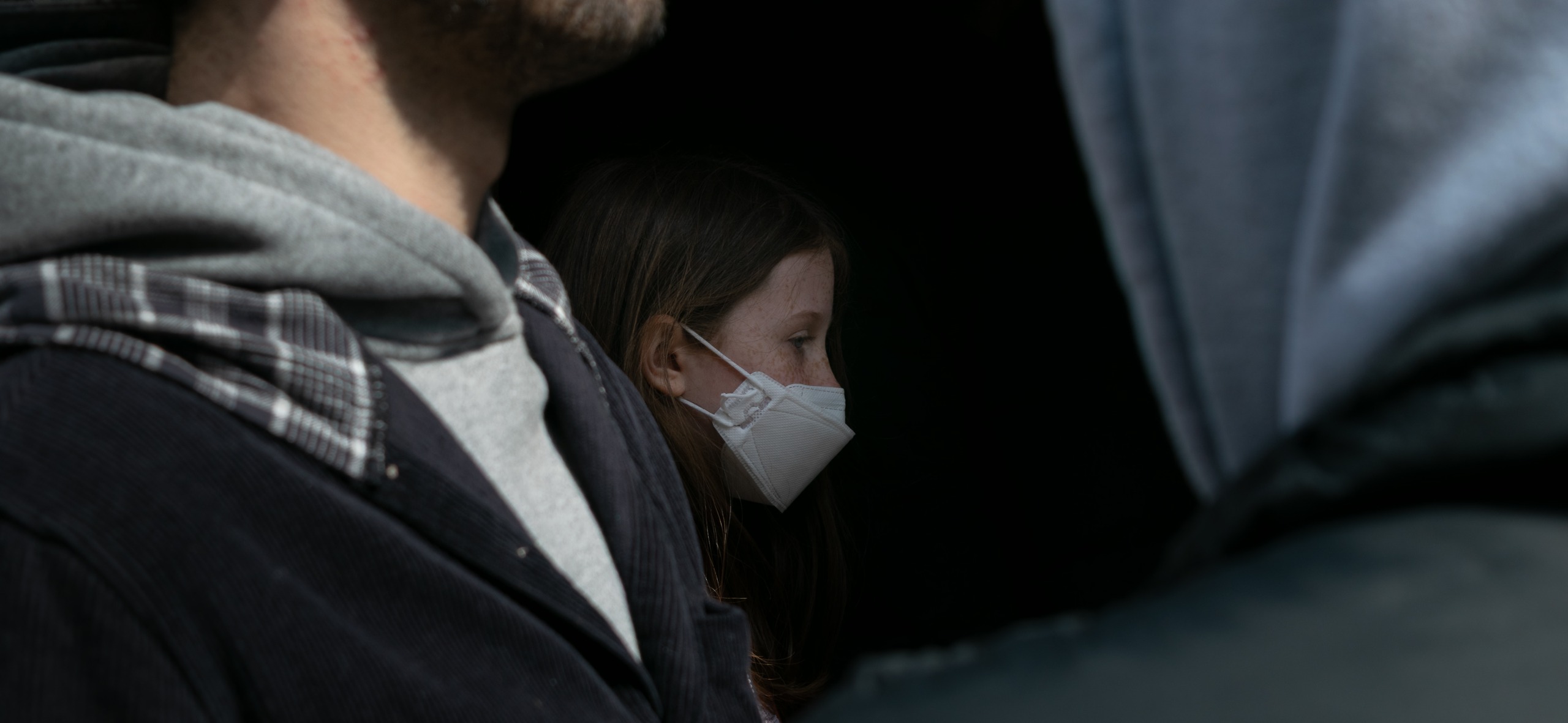 A young girl in a white KN95 mask stands in a narrow gap between two adults seen from behind, one in a gray hooded sweatshirt under a dark corduroy jacket with a plaid scarf. Her freckled face is lit in profile as she looks past the crowd into shadow.