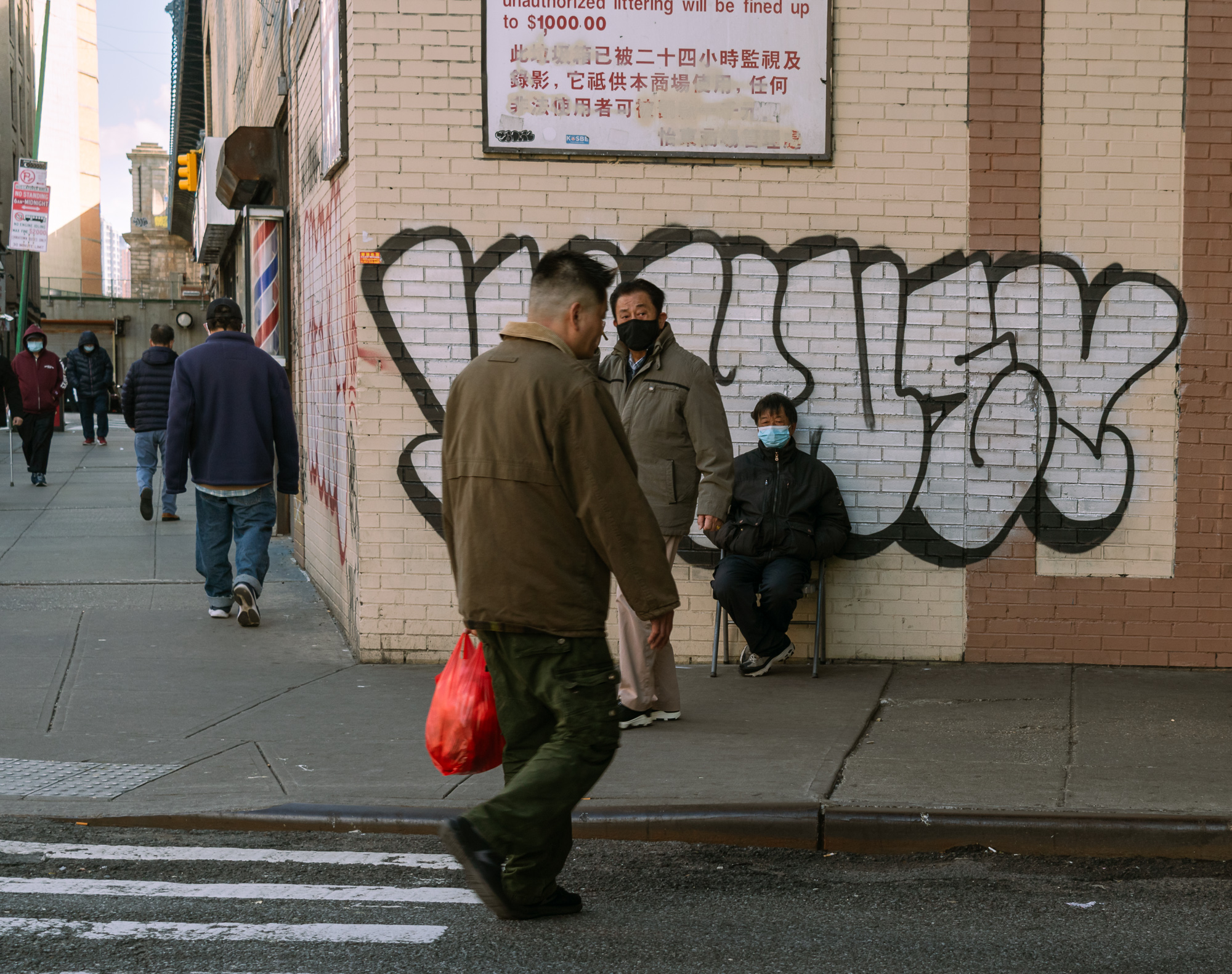 A man in an olive jacket and cargo pants carries a red plastic shopping bag as he crosses a street corner in Manhattan's Chinatown, glancing sideways. Behind him, two masked men stand and sit against a cream brick wall covered in large black graffiti tags, beneath a sign warning that unauthorized littering will be fined up to $1,000 in English and Chinese.