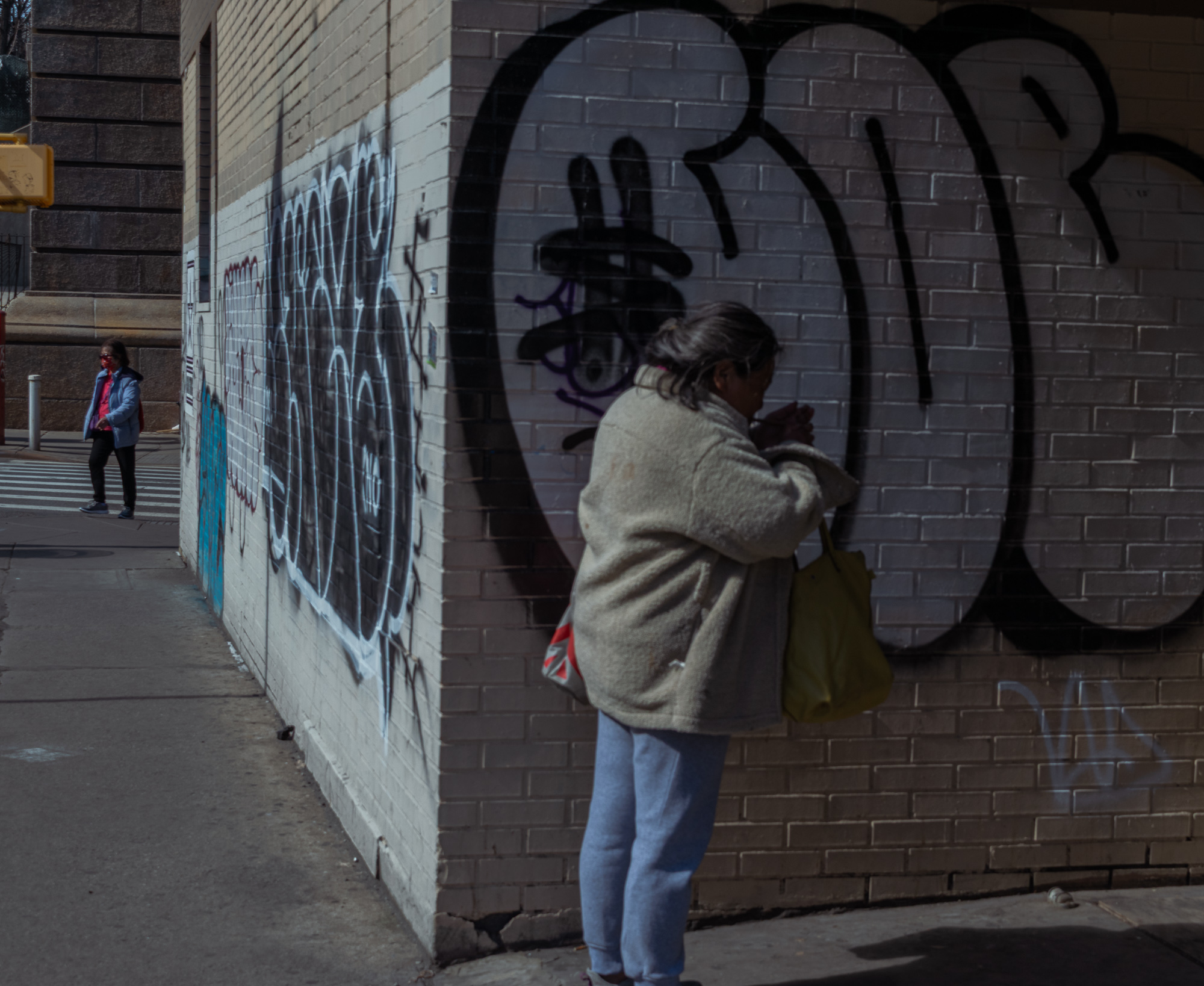 A woman in a cream fleece coat and grey sweatpants stands close to a brick wall covered in large black graffiti letters, a yellow-green tote bag on her arm as she looks down at her hands. Around the corner in bright sunlight, a second woman in a blue puffer jacket and red face mask waits at a crosswalk.