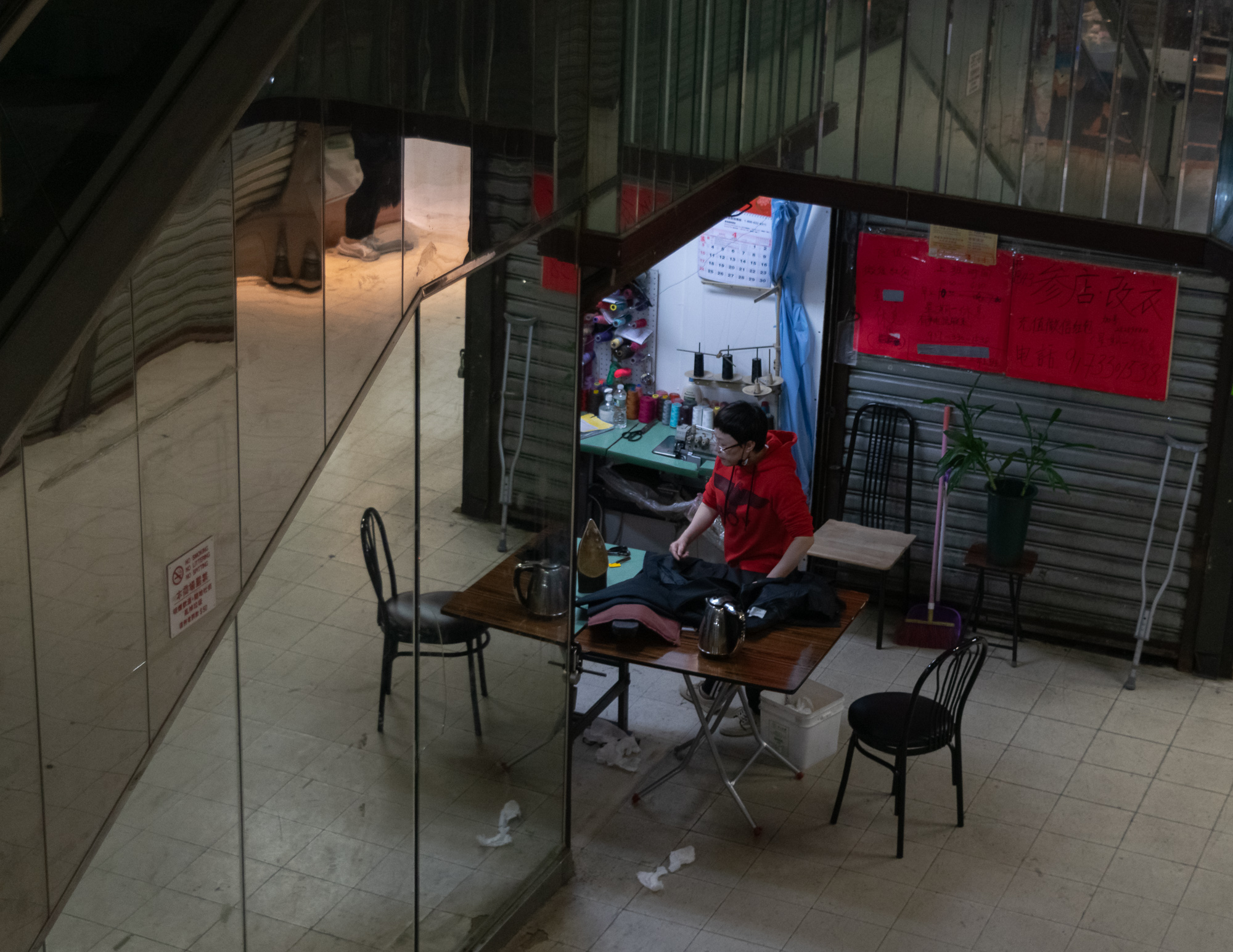 Seen from above, a man in a red hoodie stands at a folding table pressing a dark jacket inside a small alterations stall built into the space beneath a staircase, with a steam iron, metal kettles, spools of thread and a red hand-lettered Chinese sign behind him. To the left, a mirrored staircase wall reflects the legs and sneakers of someone walking on the level above.