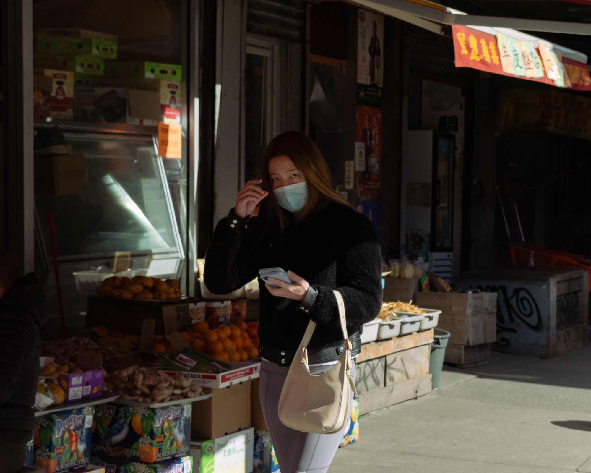 A woman in a black shearling jacket and light gray leggings stands in a patch of sunlight outside a produce shop, wearing a surgical mask and holding a white phone while tucking her hair behind her ear. Behind her are crates of oranges, loose ginger root, Blue Jay citrus boxes, and a red Chinese-character banner hanging from the awning.