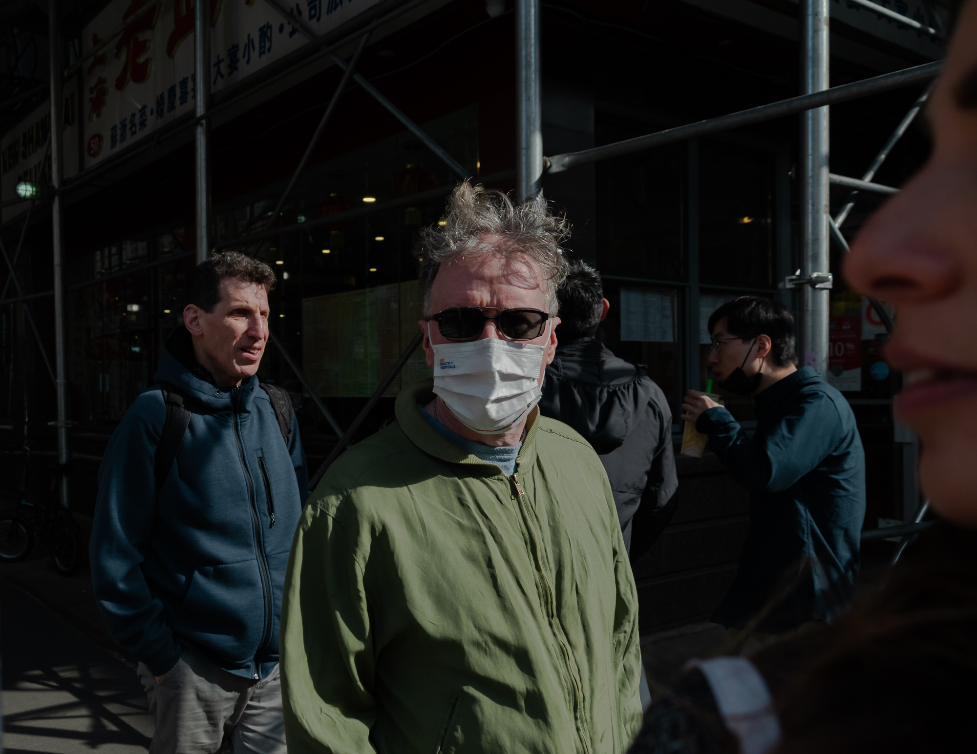 A gray-haired man in dark sunglasses, a white surgical mask and an olive green zip jacket stares directly at the camera in a shaft of hard sunlight, with a man in a blue fleece to his left, two other pedestrians behind, and Chinese-language restaurant signage and metal scaffolding poles above.