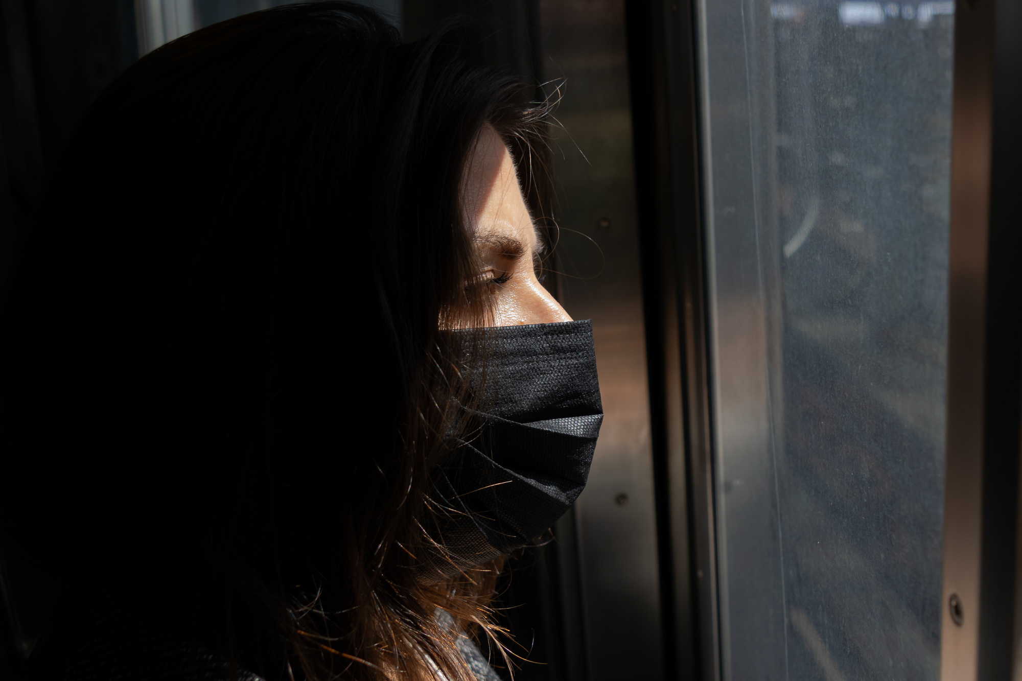 A woman in profile wearing a black pleated face mask, eyes closed, with a narrow band of sunlight crossing her brow, cheek and nose while her dark hair falls into deep shadow. Behind her is a train door frame and a scratched glass window.