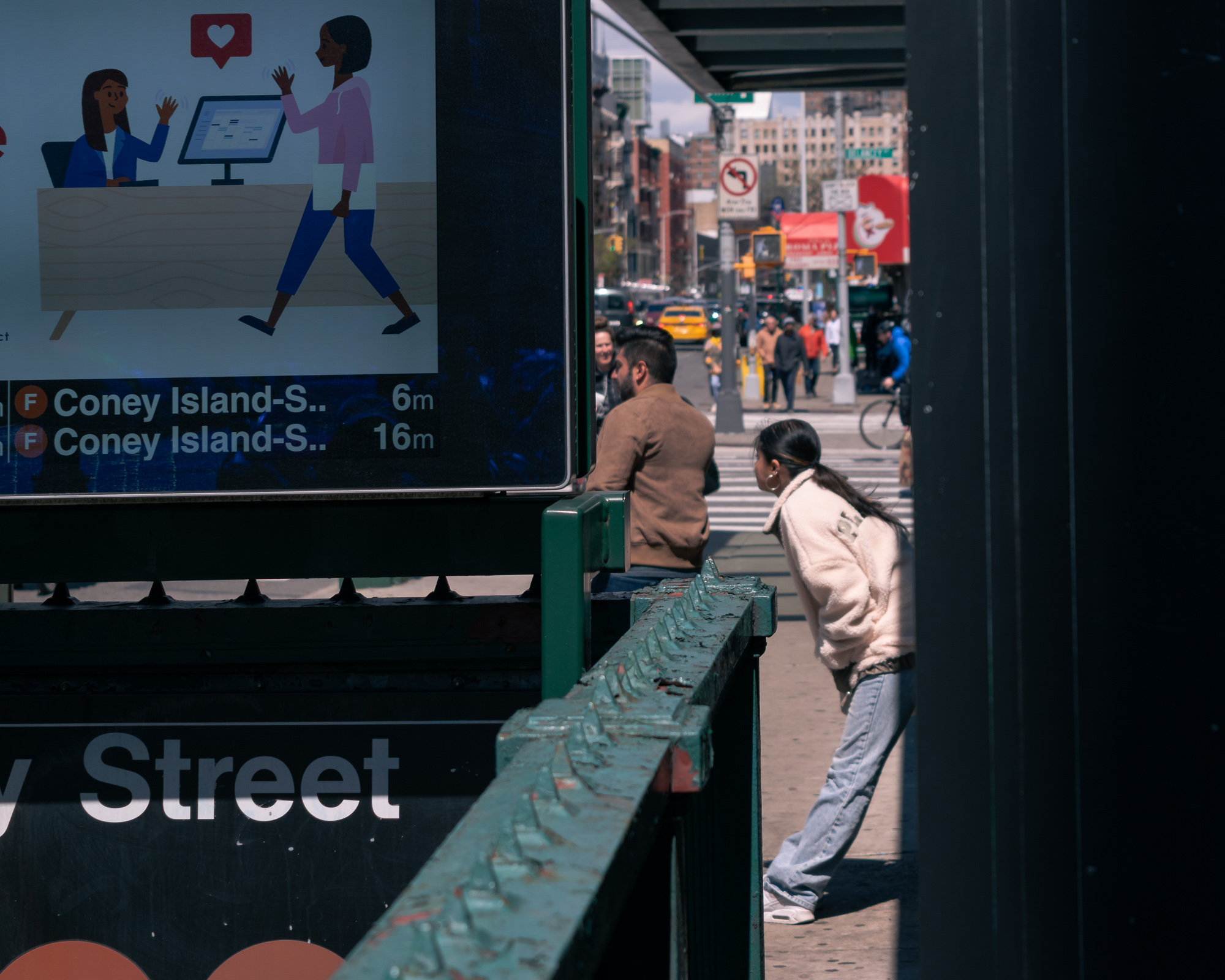 Seen through a gap beside a subway entrance, a young woman in a cream fleece jacket leans forward past a weathered green railing, looking down the sidewalk. A lit MTA screen on the left shows an illustrated ad and arrival times for two F trains to Coney Island in 6 and 16 minutes, with a crosswalk, traffic light and blurred pedestrians beyond.