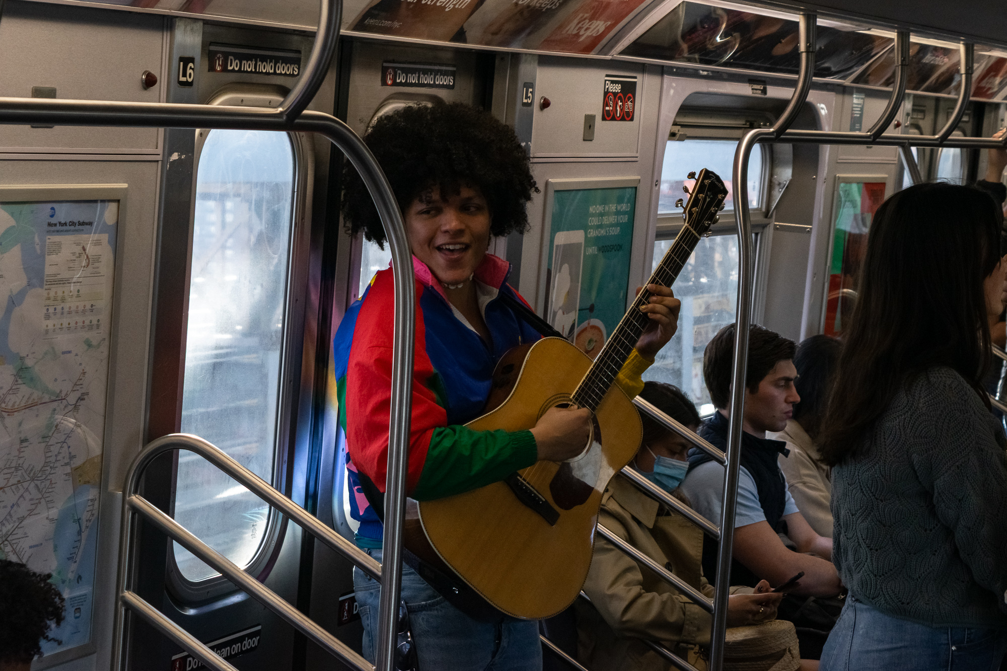 A young man with a large afro, wearing a bright multicolored windbreaker in red, blue, green and pink, stands in a crowded subway car playing an acoustic guitar and singing. Behind him a door sign reads 'Do not hold doors' and seated passengers, one in a face mask, look down at phones.