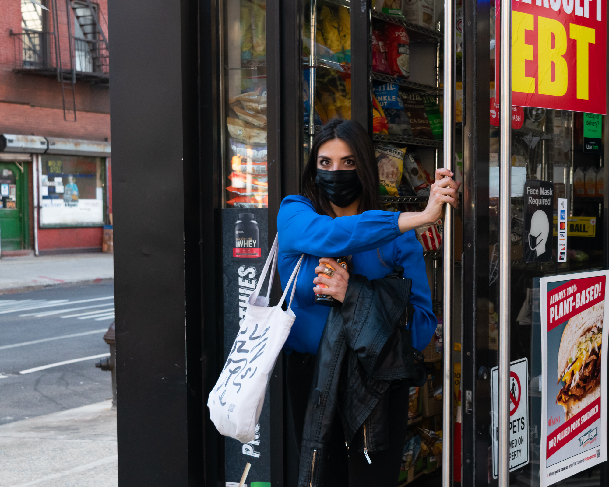 A woman in a black face mask and bright blue blouse pushes open the glass door of a corner bodega, holding a bottle and a black leather jacket against her chest while a white canvas tote hangs from her arm. Behind her are shelves of chip bags, and the door glass carries a red EBT sign, a Face Mask Required notice, and a plant-based sandwich poster.