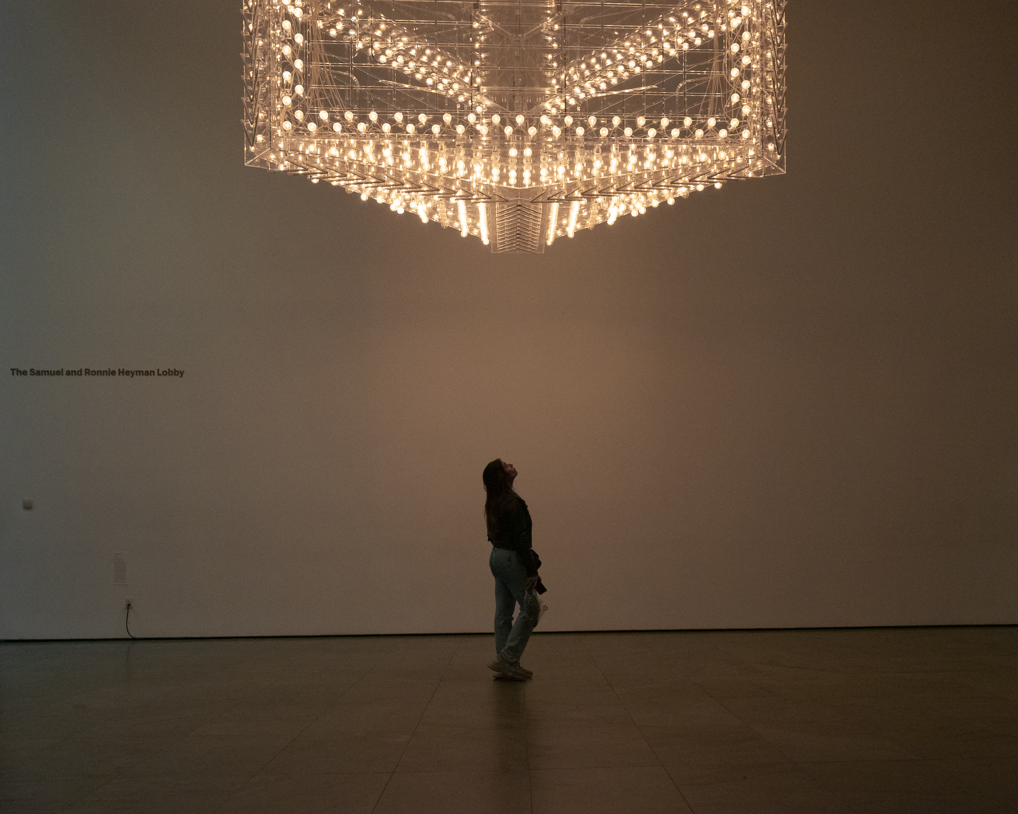 A woman stands alone in a dim museum lobby, silhouetted against a pale wall as she tilts her head back to look up at a massive suspended sculpture made of hundreds of lit bulbs on a clear tiered frame. Small text on the wall to her left reads The Samuel and Ronnie Heyman Lobby.