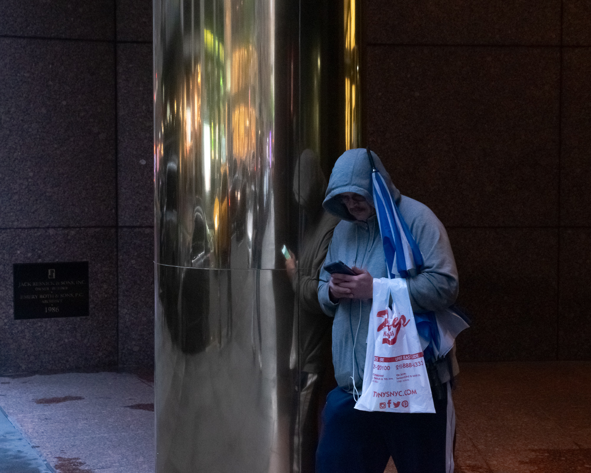 A man in a grey hooded sweatshirt leans against a polished steel column outside a granite office building, looking down at a phone in both hands with white earbud cords trailing. A folded blue umbrella hangs over his shoulder and a white plastic takeout bag dangles from his arm; his reflection, warped and doubled, stretches across the column beside him.