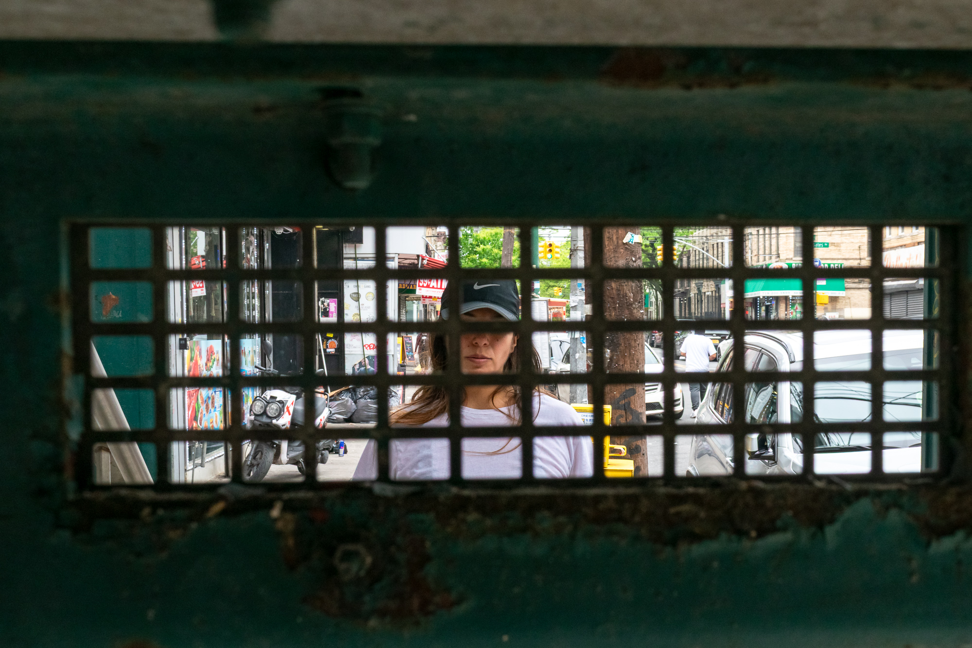 Seen through a small barred vent in a weathered green metal wall, a woman in a black Nike cap and white long-sleeve shirt stands on a Brooklyn sidewalk. Behind her are a parked white scooter, black garbage bags, bodega storefronts, parked cars and a Gates Ave street sign.
