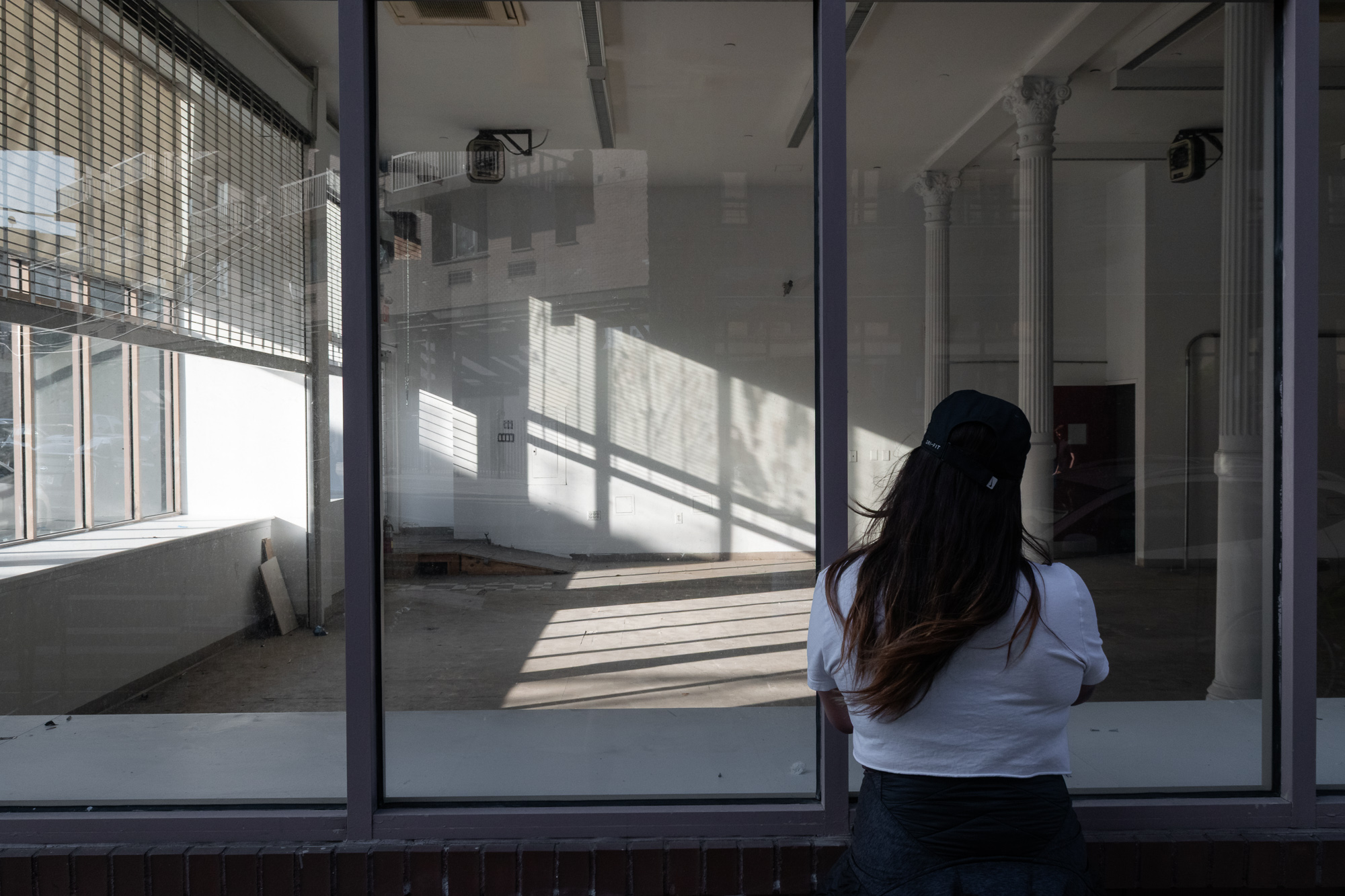 A woman seen from behind in a white t-shirt and black baseball cap leans against the glass of a vacant ground-floor storefront, looking into a gutted interior where late afternoon sun casts long diagonal window-frame shadows across the bare concrete floor. The glass doubles as a mirror, reflecting the brick building and fire escape across the street.