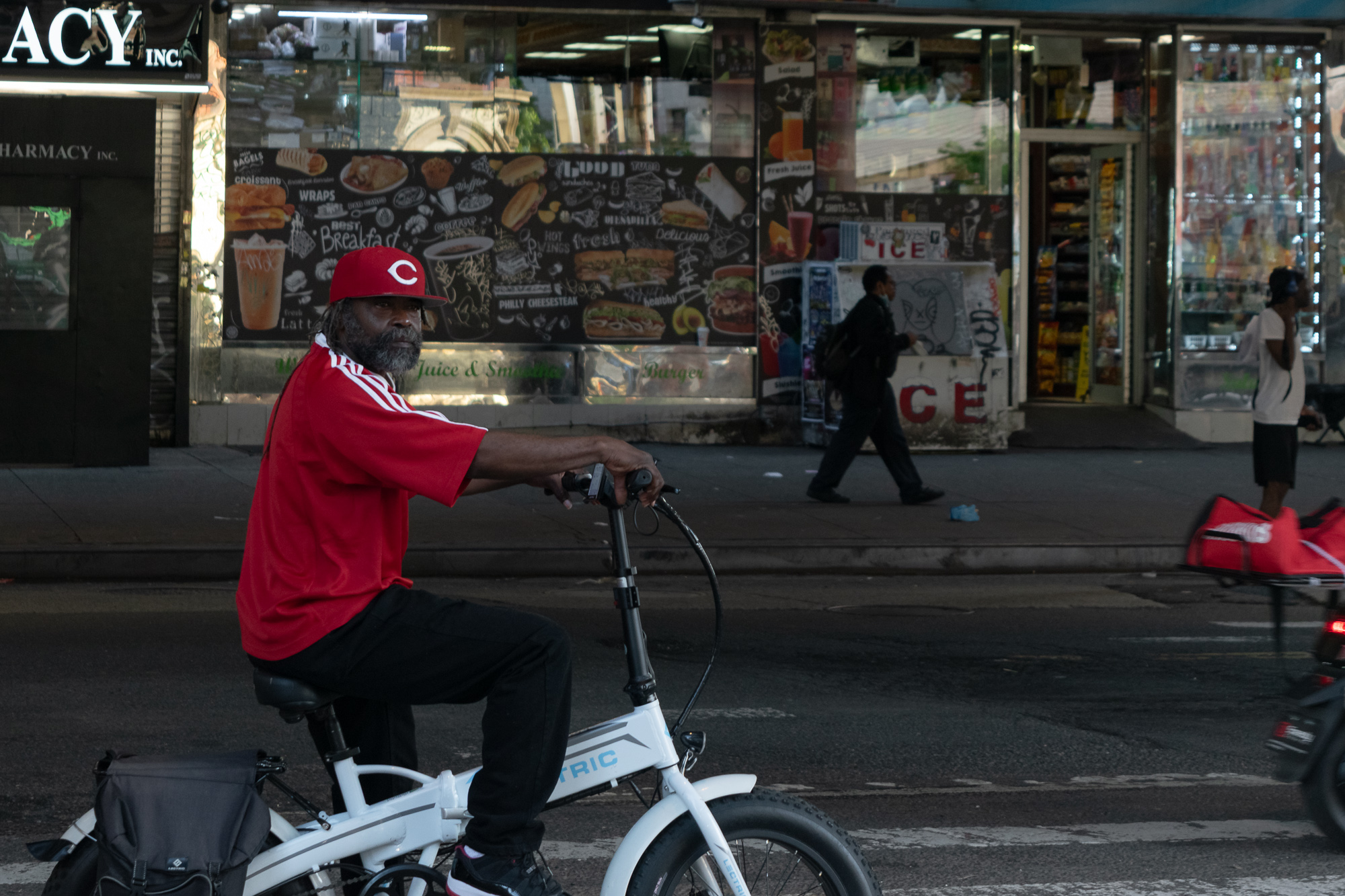 A bearded man in a red Cincinnati 'C' cap and red striped jersey sits astride a white folding electric bike in the street, turning to look back at the camera. Behind him a bodega window is covered with a chalk-style menu board advertising wraps, Philly cheesesteak and fresh juice, with an ICE cooler and a pharmacy shutter further down the block.
