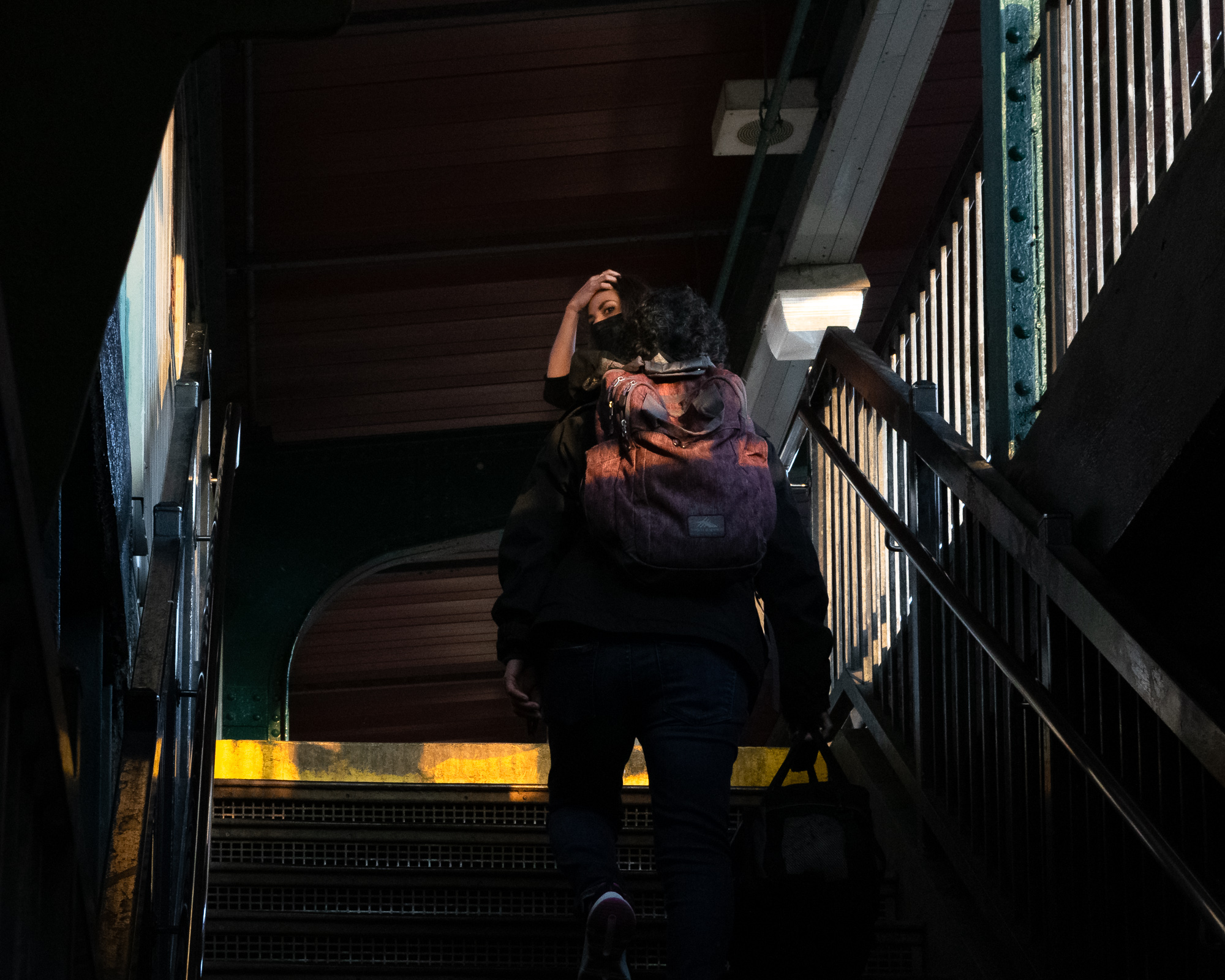 Seen from below on a dim elevated subway staircase, a person in a dark jacket with a faded pink and purple backpack climbs toward a landing. Ahead of them a woman in a black face mask pushes her hair back and turns to look back over her shoulder, lit by a small fluorescent fixture against red wooden walls.