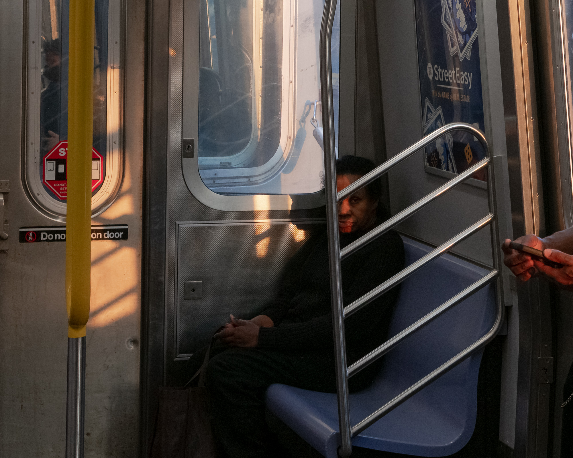 Inside a New York subway car, a woman in a black sweater sits on a blue bench seat beside the doors, her face lit by a narrow band of orange evening light while chrome handrails cut diagonally across the frame. At right, a hand holds a phone near the door; a yellow pole and a StreetEasy ad fill the left and upper edges.