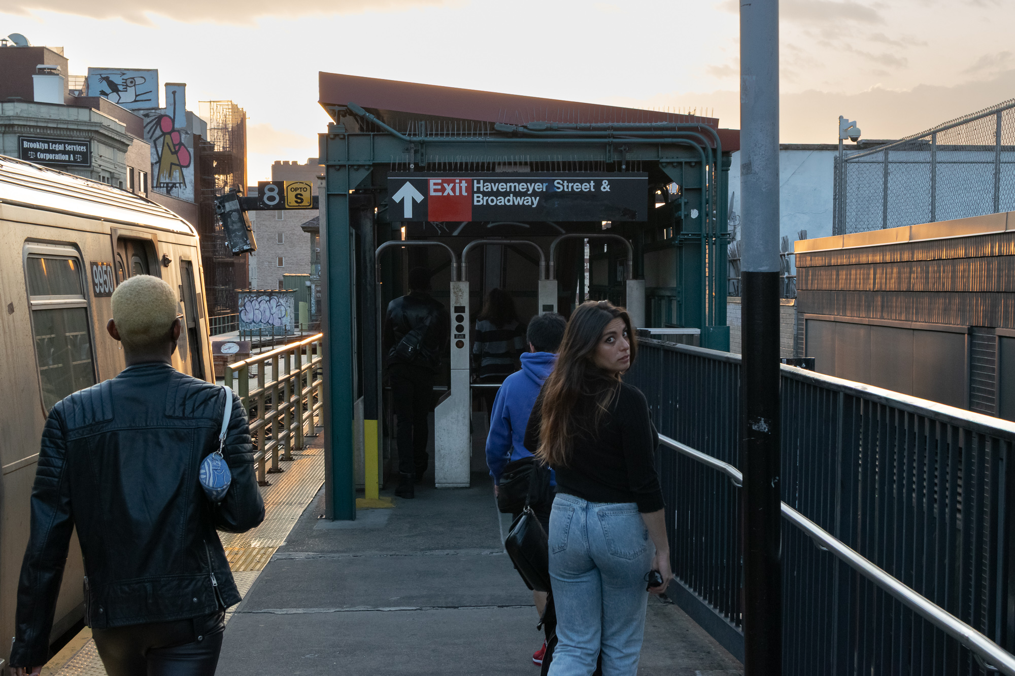 Commuters walk along an elevated subway platform toward a covered stairway marked with a black and red sign reading Exit, Havemeyer Street & Broadway. A woman in a black top and light jeans turns to look back over her shoulder, while a man with bleached blond hair and a leather jacket walks ahead near a waiting silver train.