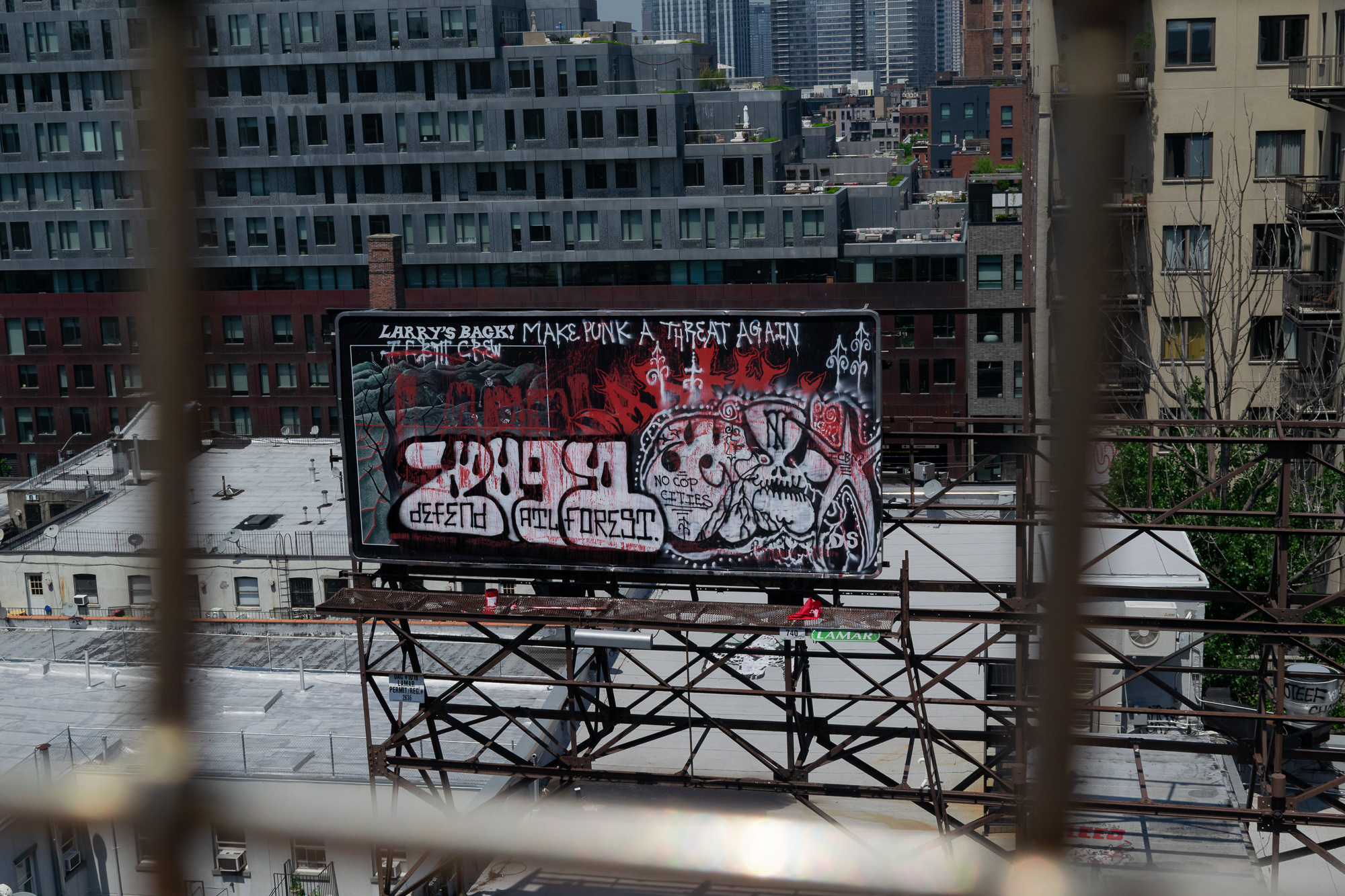 Seen through the bars of a fire escape or window guard, a large Lamar billboard on a rusted steel catwalk has been painted over entirely with red, white and black graffiti reading 'Larry's Back! Make Punk a Threat Again,' 'Defend ATL Forest' and 'No Cop Cities,' with a skull drawing at right. Behind it rise gray apartment blocks, tar rooftops and distant towers.