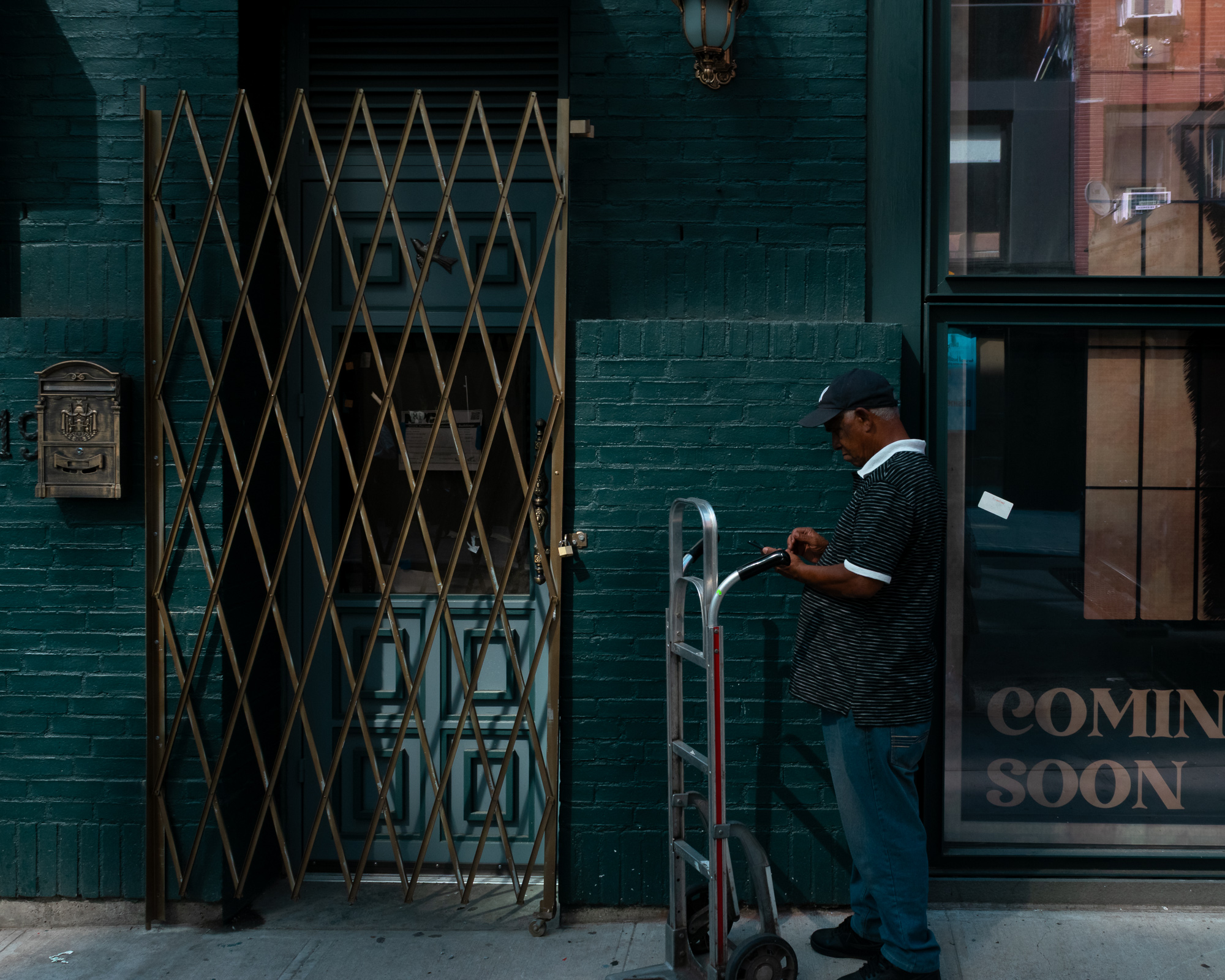 A man in a navy baseball cap and black and white striped polo shirt stands beside an aluminum hand truck, looking down at a phone in his hands. Behind him is a dark green painted brick wall with a brass accordion security gate, a brass mailbox, and a shop window with COMING SOON lettering.