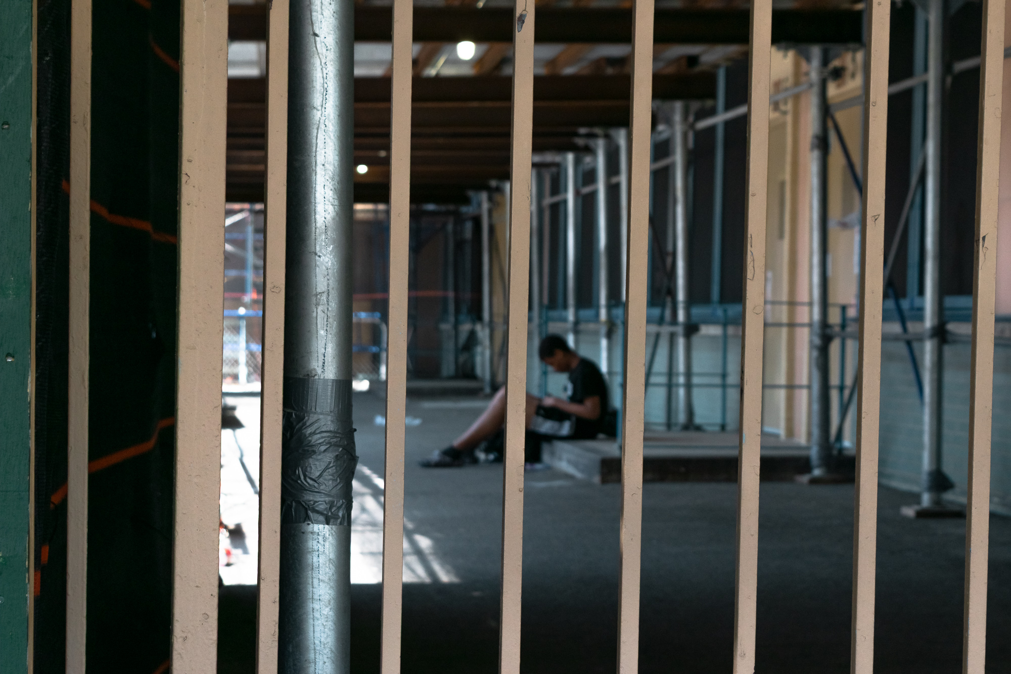 Looking through a row of vertical white railing bars at a shadowed construction passage, where a person in a black t-shirt and shorts sits on a low concrete step, head down, hands in their lap. A galvanized scaffold pipe wrapped in black duct tape runs down the left of the frame, with scaffolding uprights and a bare ceiling light behind.