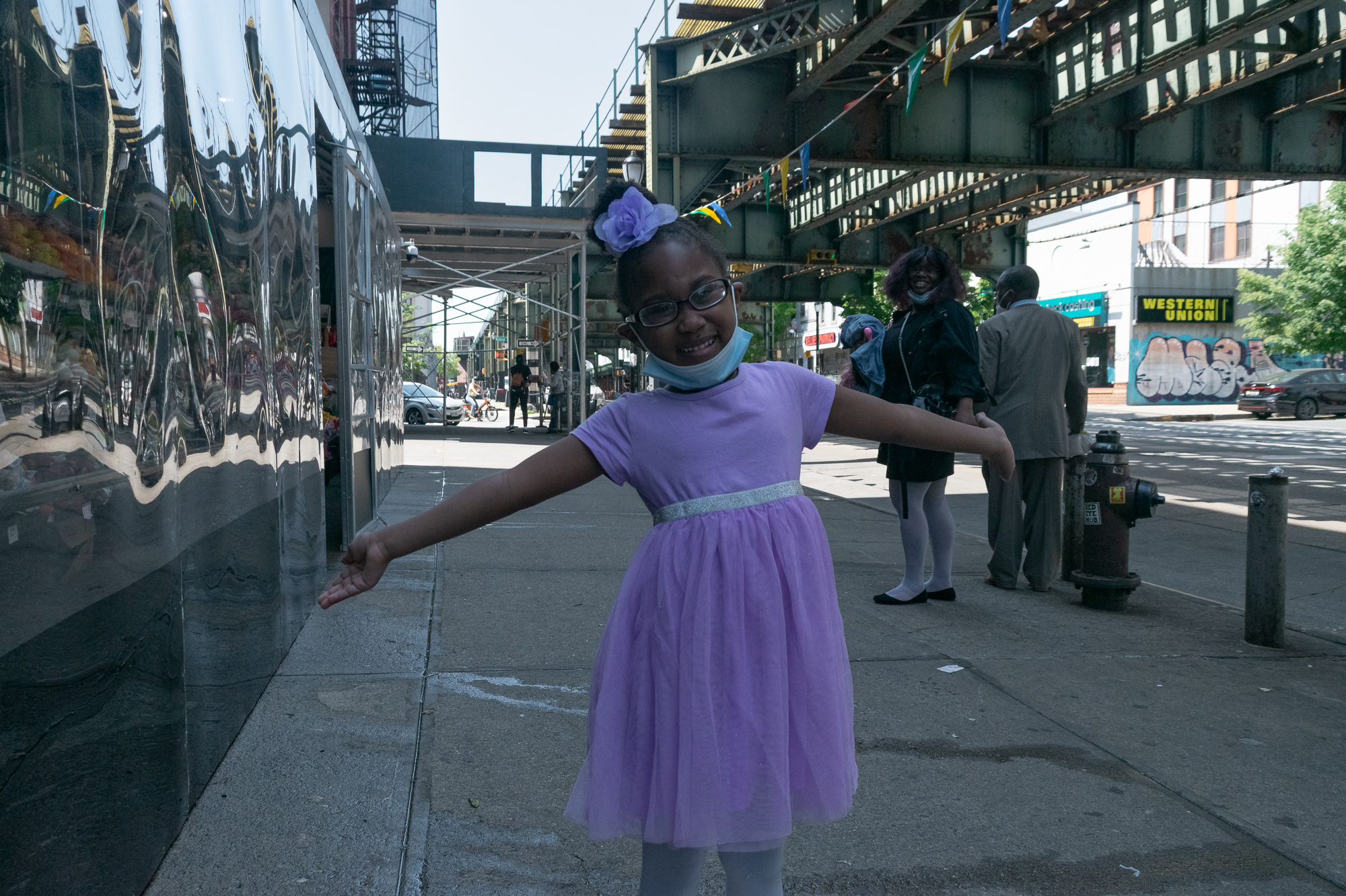 A young girl in a lavender tulle dress and glasses stands on a sidewalk with her arms stretched wide, a surgical mask pulled down under her chin and a purple flower clip in her hair. Behind her, an elevated train structure crosses the street, with a woman in black, a man in a tan suit, a fire hydrant and a Western Union sign in the background.
