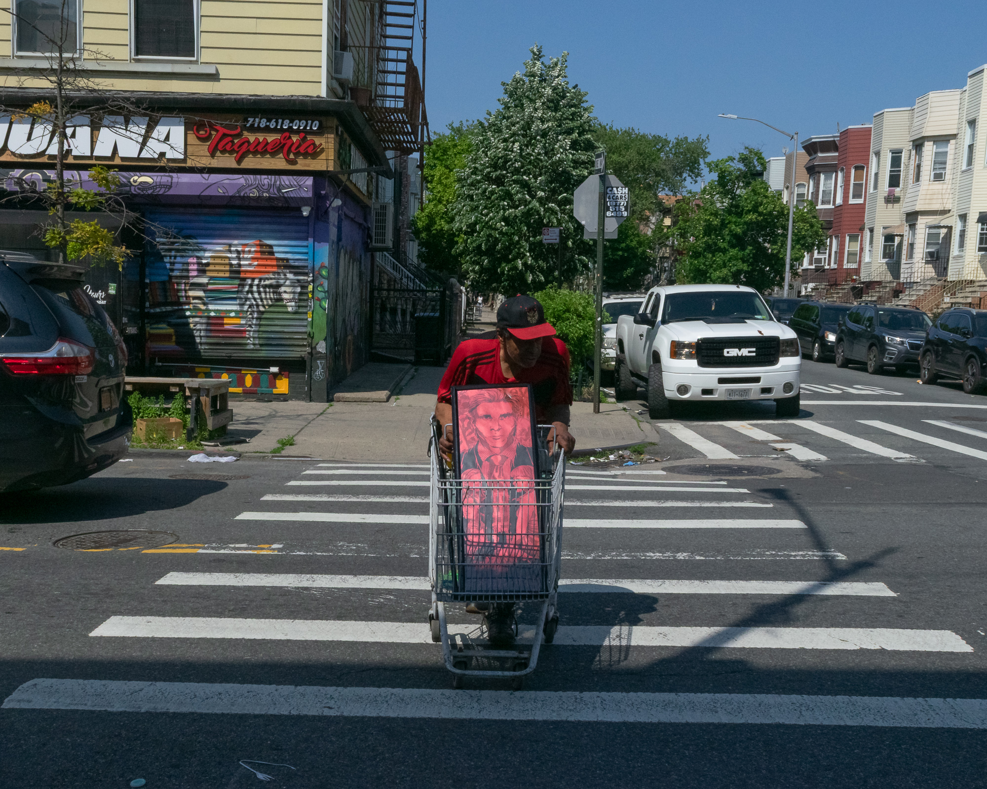 A man in a red cap and red track jacket pushes a shopping cart holding a large framed portrait printed in bright red across a marked crosswalk, with a corner taqueria and graffiti-covered roll gate behind him and a white GMC pickup parked at the curb.