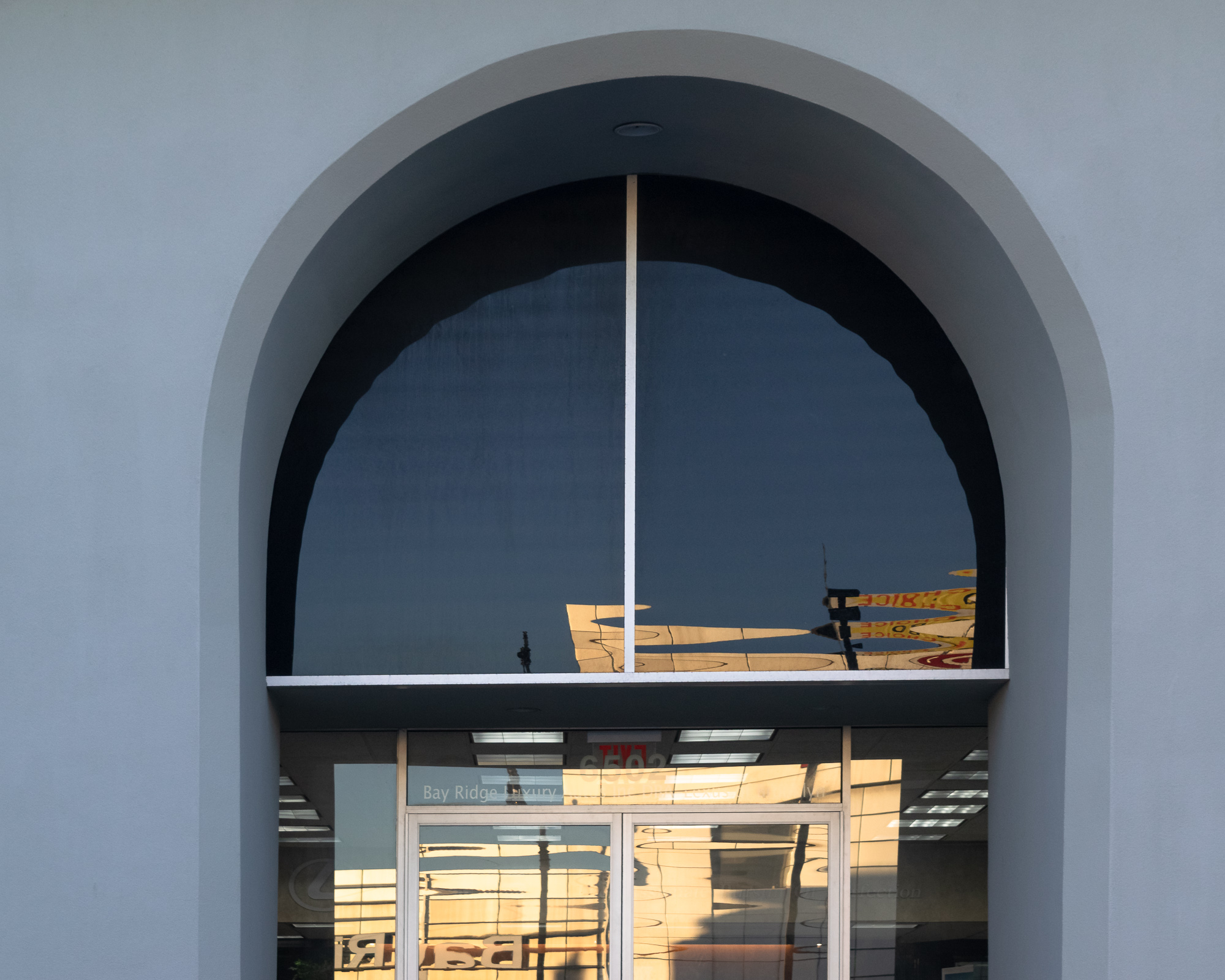 A large arched window set into a pale gray stucco wall, its dark tinted glass reflecting a sunlit yellow awning, a traffic signal and utility pole. Below the transom, glass doors read 'Bay Ridge Luxury' with fluorescent ceiling lights and a Lexus logo visible inside the showroom.