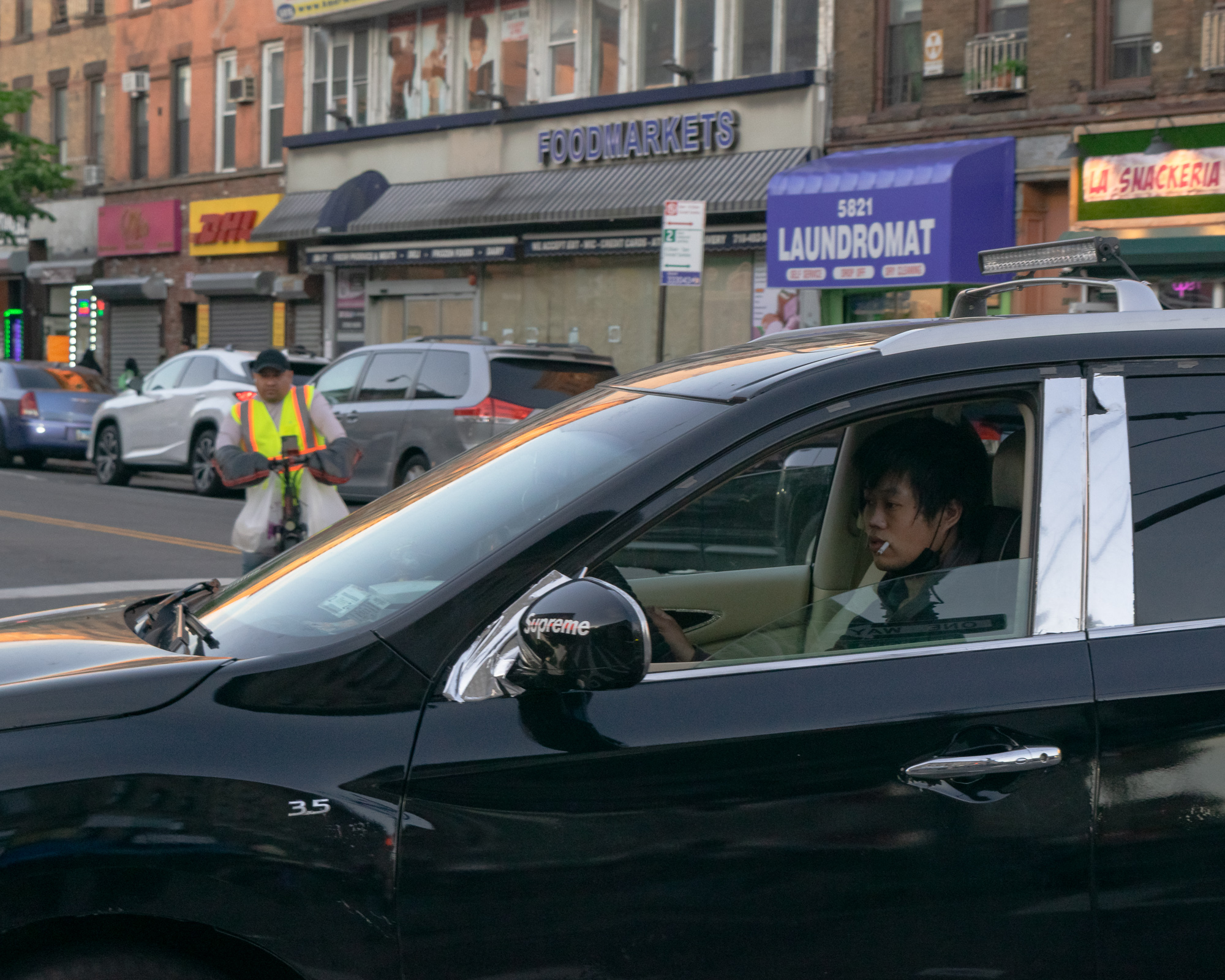 A young man sits behind the wheel of a black SUV with an unlit cigarette in his mouth, one hand on the door near a chrome side mirror wrapped in Supreme tape. Behind the car, a storefront row shows a Foodmarkets sign, a DHL awning, a purple 5821 Laundromat sign, and a man in a yellow safety vest standing with a bike.