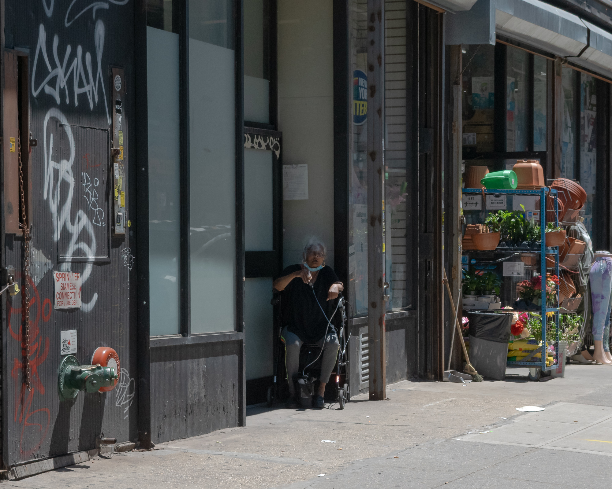 An older woman in black sits with a rolling walker in the deep shade of a recessed storefront doorway, a surgical mask pulled down under her chin. To her left a graffiti-covered metal roll gate carries a Siamese sprinkler connection sign and a green fire hydrant standpipe; to her right, a sunlit garden shop displays terracotta pots, flowers and plants on blue steel shelving.
