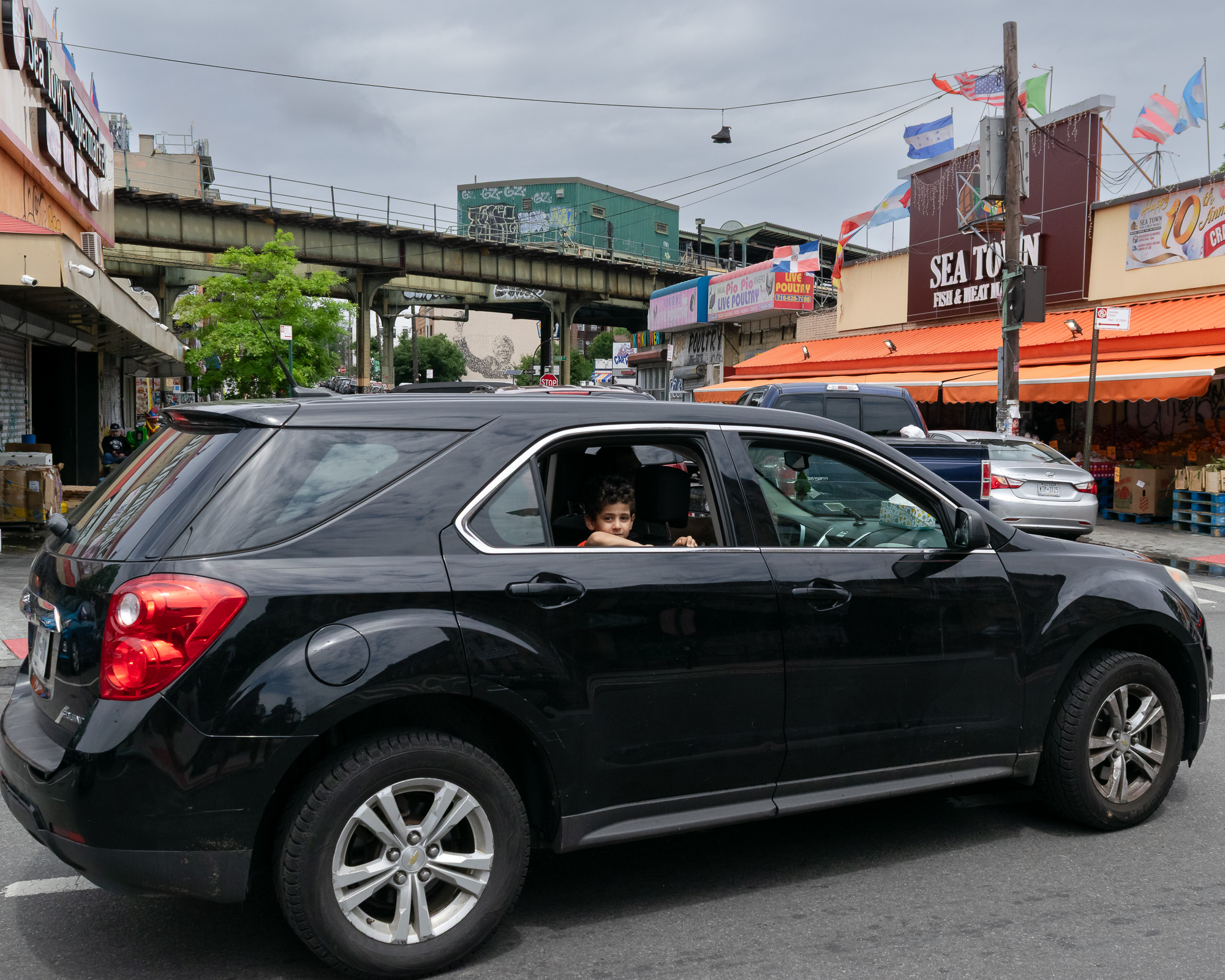 A boy rests his arms on the open rear window of a black Chevrolet SUV stopped in a city street, looking directly at the camera. Behind him are an elevated subway viaduct, a Sea Town Fish & Meat Market with an orange awning, a Live Poultry sign, and rows of flags on a utility pole.