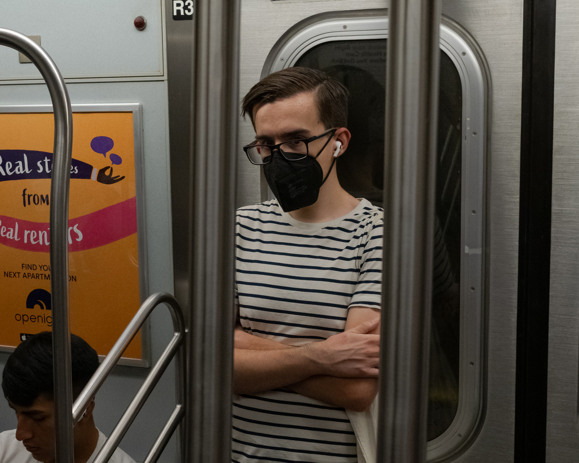 A young man in glasses, a black KN95 mask, and a navy-and-white striped t-shirt stands with arms crossed against a subway door, one white wireless earbud visible, his body framed between two vertical stainless steel poles. To the left, an orange apartment-rental ad and the door marking R3 are visible, with another passenger's head at the bottom edge of the frame.