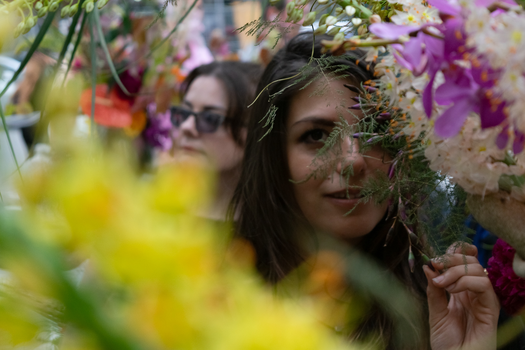 A dark-haired woman looks toward the camera through a screen of asparagus fern and pink orchids at a flower stall, her hand raised to hold a stem aside. Behind her, a second woman in sunglasses stands out of focus among the blooms, and a wash of blurred yellow flowers fills the foreground.