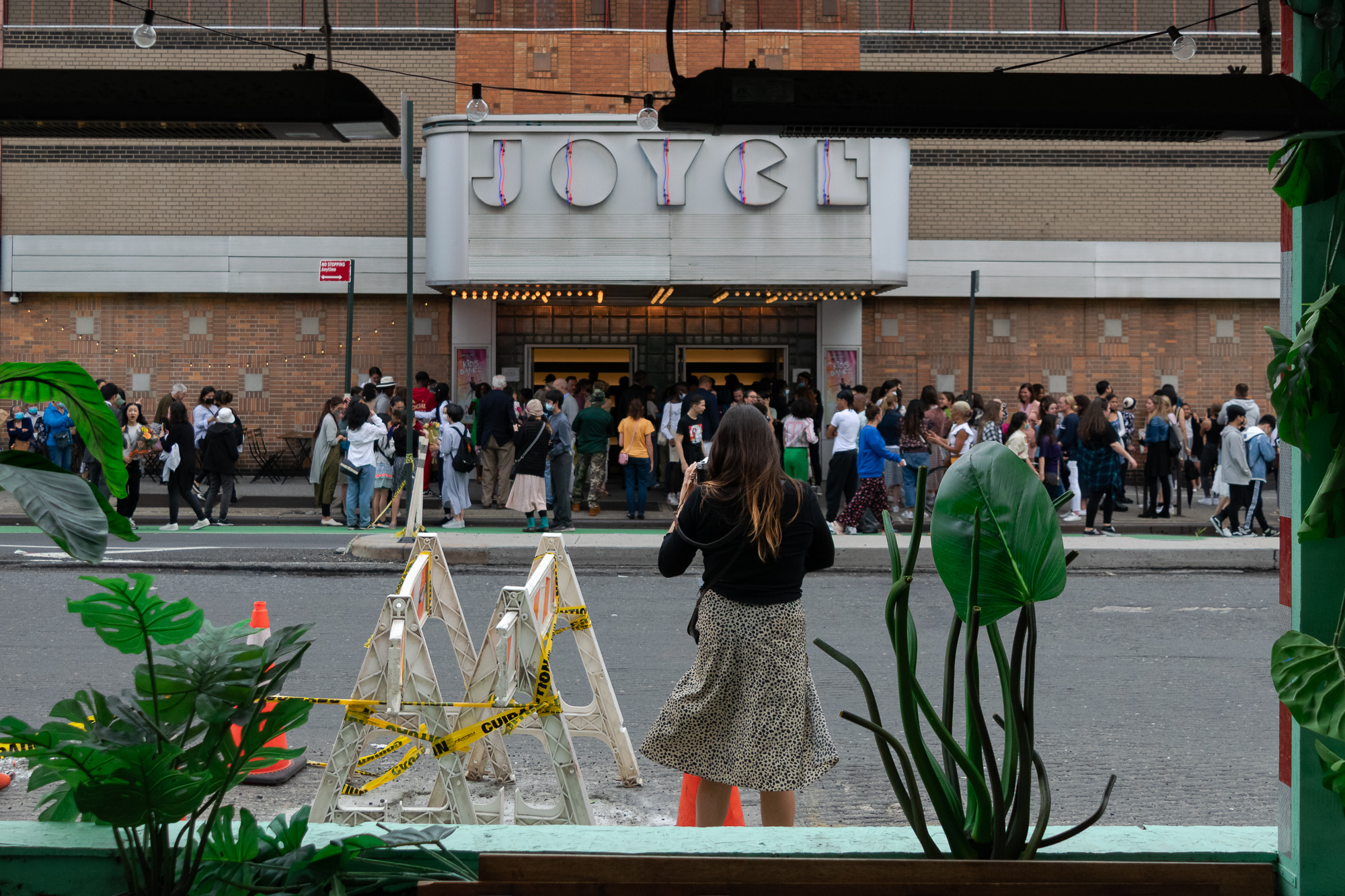 Framed by potted plants on a cafe windowsill, a woman in a black top and leopard-print skirt stands on Eighth Avenue holding a phone, facing a crowd spilling out of the Joyce Theater under its Art Deco marquee. Yellow caution tape wraps a pair of white A-frame barricades and an orange traffic cone in the street between them.
