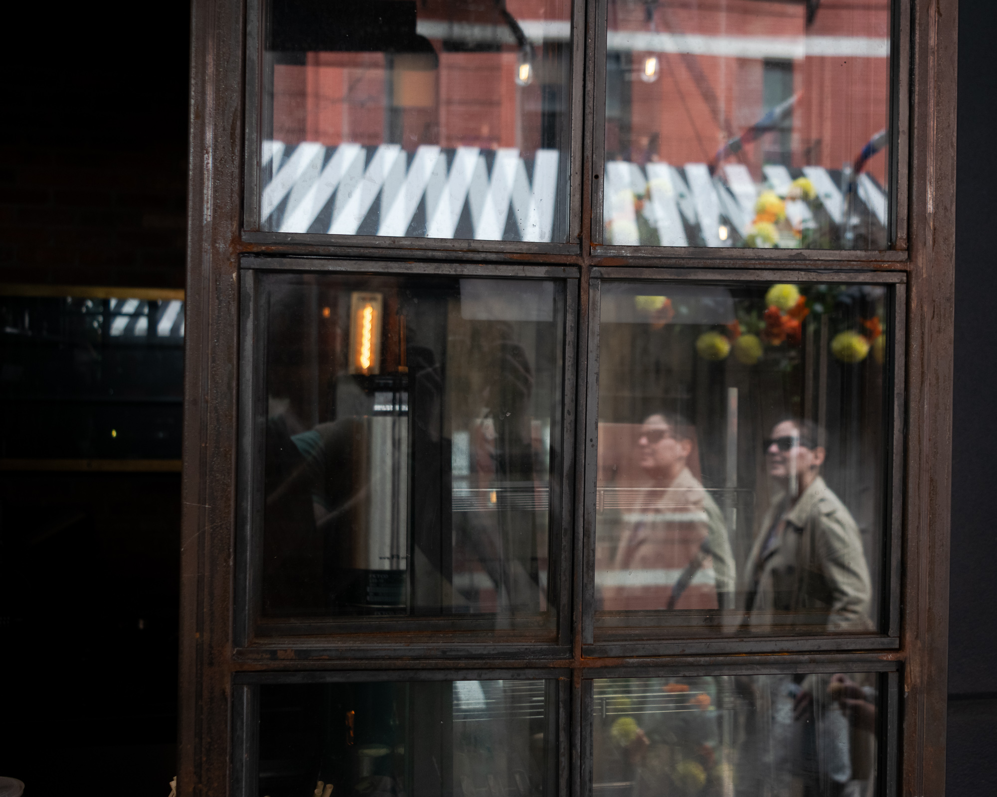 A large window divided into panes by a rusted steel frame reflects a street scene: a red brick building, a black and white striped awning, hanging orange and yellow flowers, and two people in sunglasses and tan coats walking past. Inside the dark interior, a single warm filament bulb glows behind the glass.