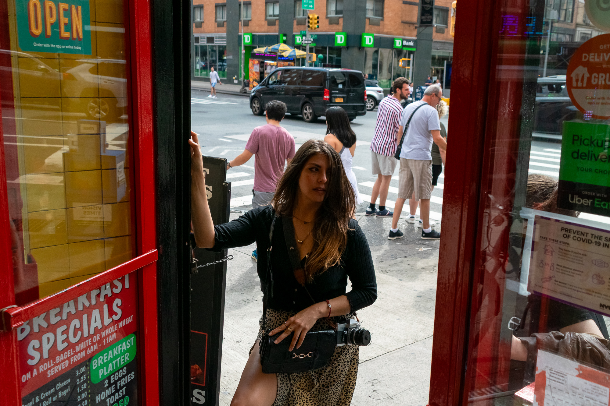 Shot from inside a deli, a woman in a black cardigan and leopard-print skirt holds the red doorframe as she steps out, a 35mm film camera hanging at her hip beside a black handbag. Behind her, pedestrians wait at a crosswalk under a TD Bank branch and a yellow-umbrella food cart.