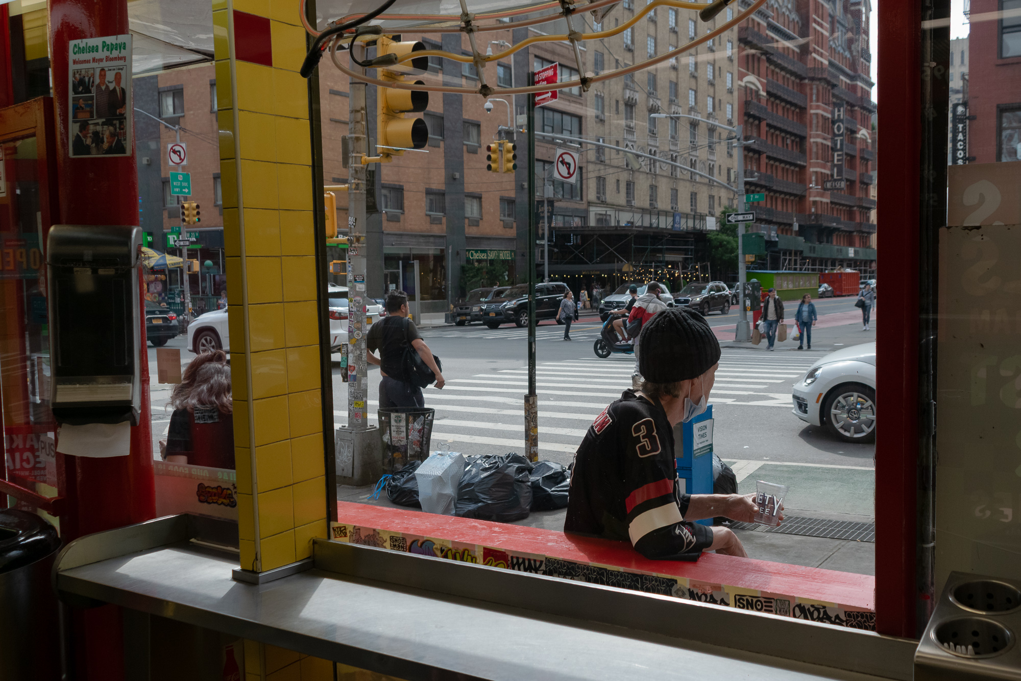 View from inside a Chelsea hot dog counter through a large window: a man in a black knit cap and a black number 3 jersey leans on the red exterior ledge holding a clear plastic cup, mask pulled below his chin, while traffic lights, trash bags, pedestrians and the Hotel Chelsea fill the avenue behind him.