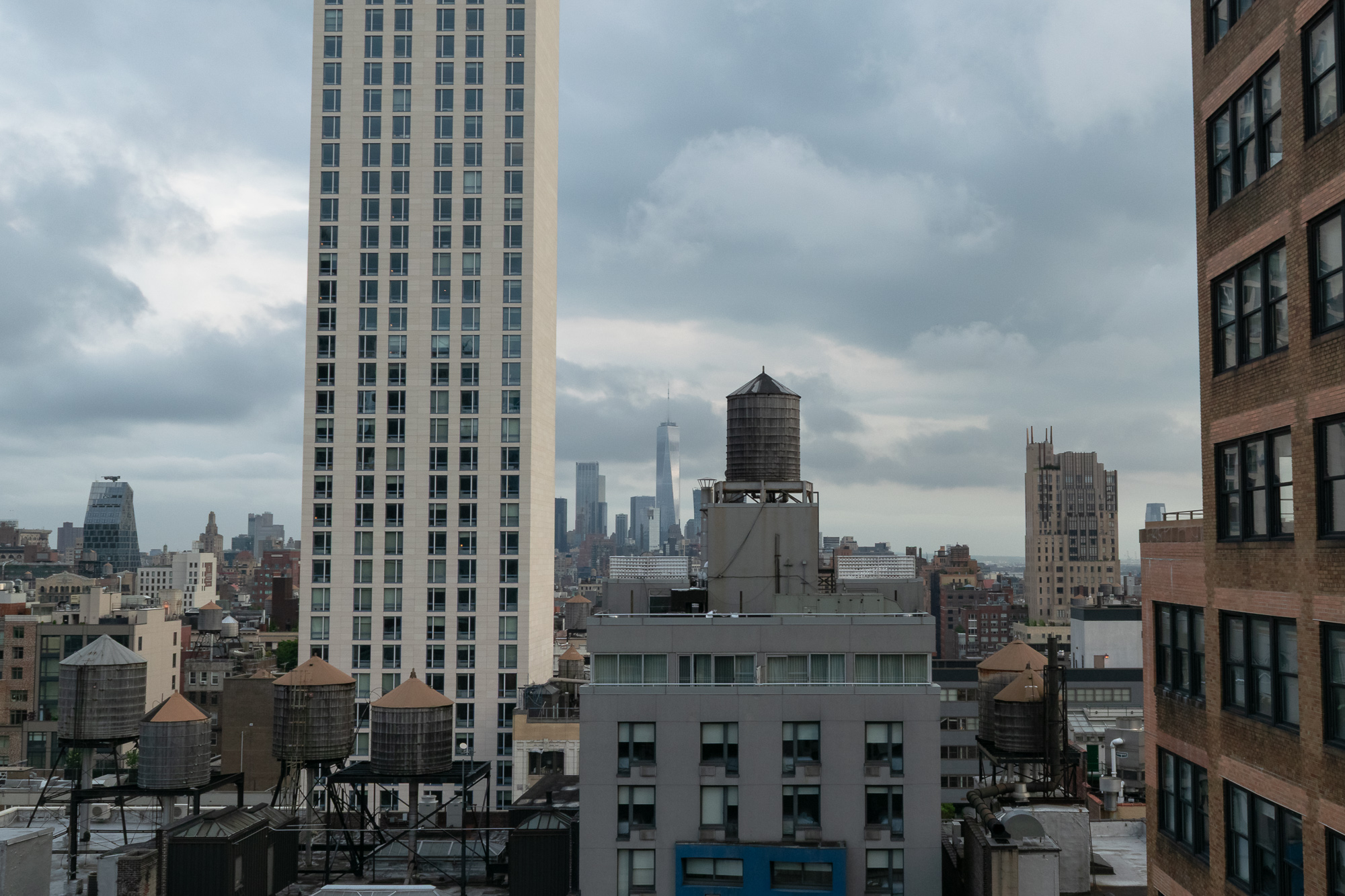 A view across Manhattan rooftops crowded with wooden water towers, with a pale high-rise tower rising at left and One World Trade Center visible in the hazy distance beneath heavy grey clouds.