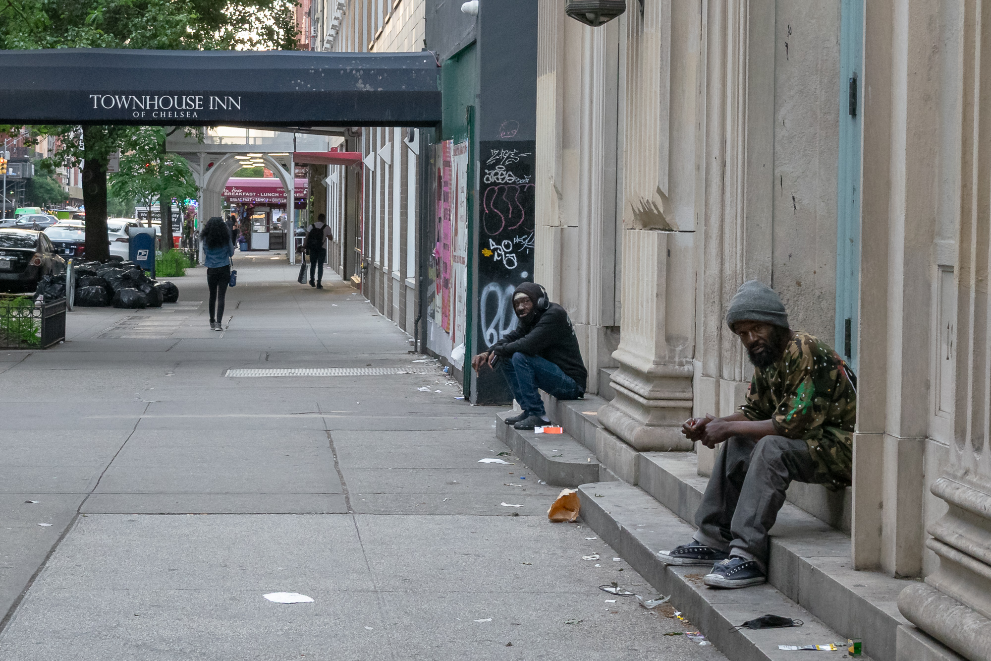 On a Chelsea sidewalk, two men sit on the stone steps of a boarded-up building: one in a dark hoodie and headphones crouched near a graffiti-tagged wall, the other in a camouflage jacket and grey knit cap looking directly at the camera. Down the block, pedestrians pass under a navy awning reading Townhouse Inn of Chelsea.