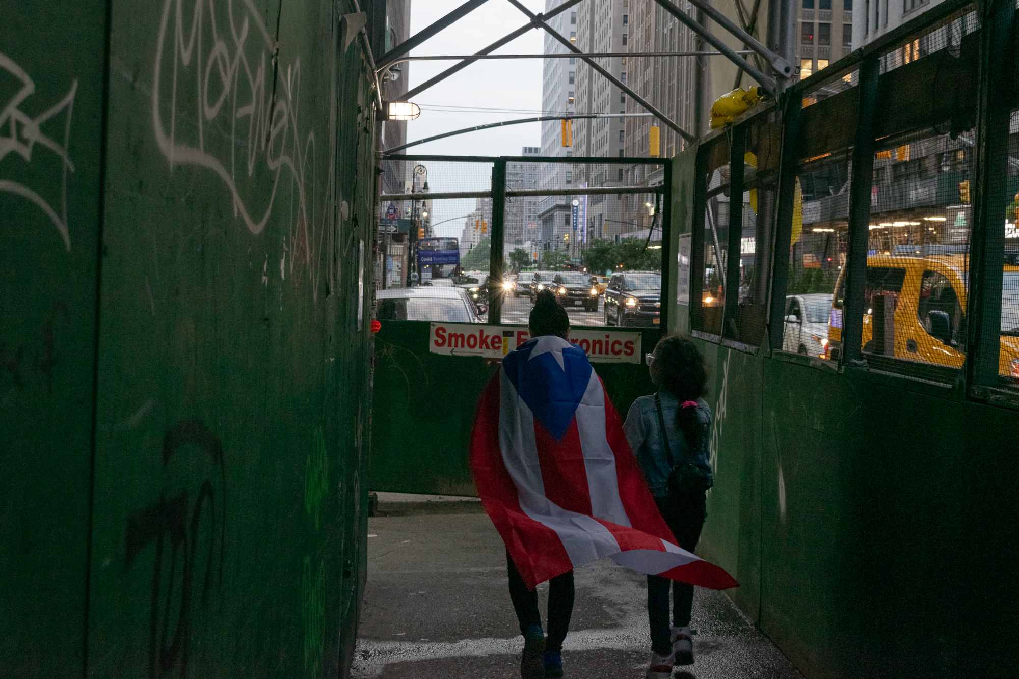 Two young people walk away from the camera through a green plywood construction walkway, one with a Puerto Rican flag draped over her shoulders like a cape. Beyond the tunnel opening, traffic moves along a Manhattan avenue past a red and white 'Smoke Shop Electronics' sign and a yellow truck.