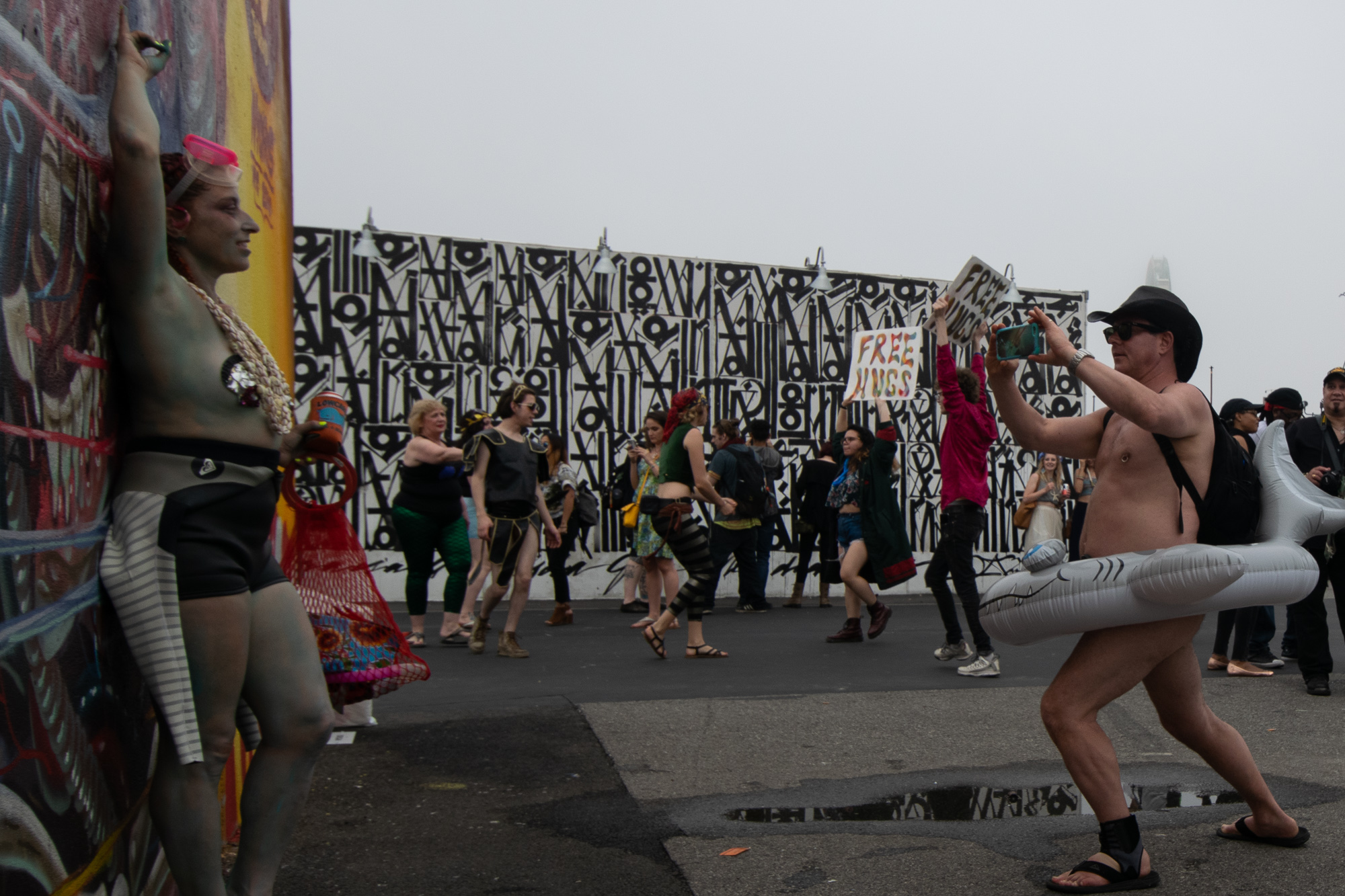 A naked man wearing a black cowboy hat, sunglasses and sandals leans forward to film with a phone while an inflatable shark float hangs around his waist; across the street a silver body-painted person in swim goggles and shorts poses against a graffiti wall as a crowd passes a black-and-white mural and a hand-lettered FREE HUGS sign.