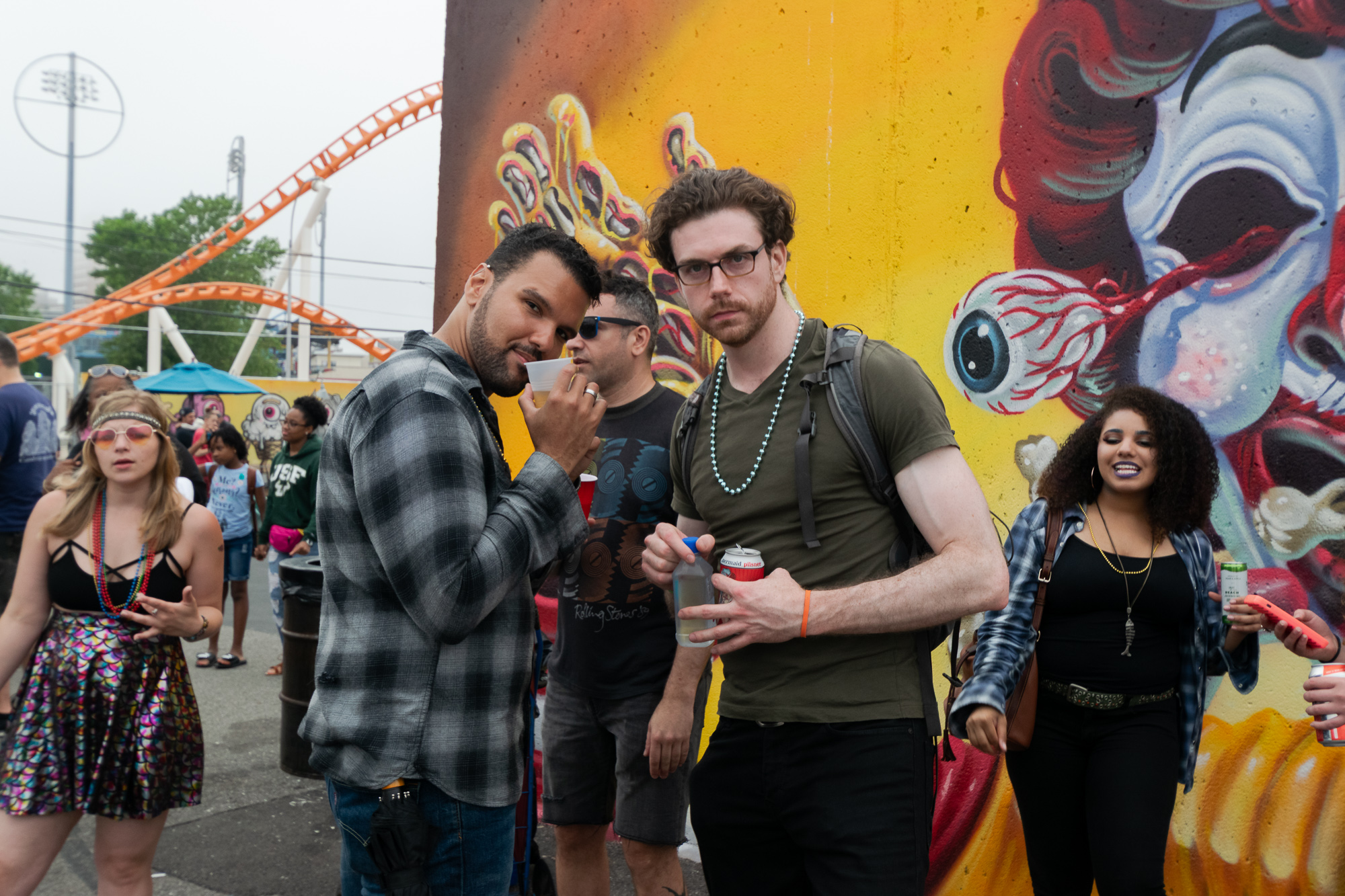 Two men stand close to the camera in front of a large wall mural of a red-haired clown face with a bulging eyeball, one in a gray plaid shirt sipping from a plastic cup, the other in an olive T-shirt and beaded necklace holding a can of pilsner and a clear water bottle. Behind them a crowd fills a paved lot, with the orange track of a roller coaster rising against a flat white sky.