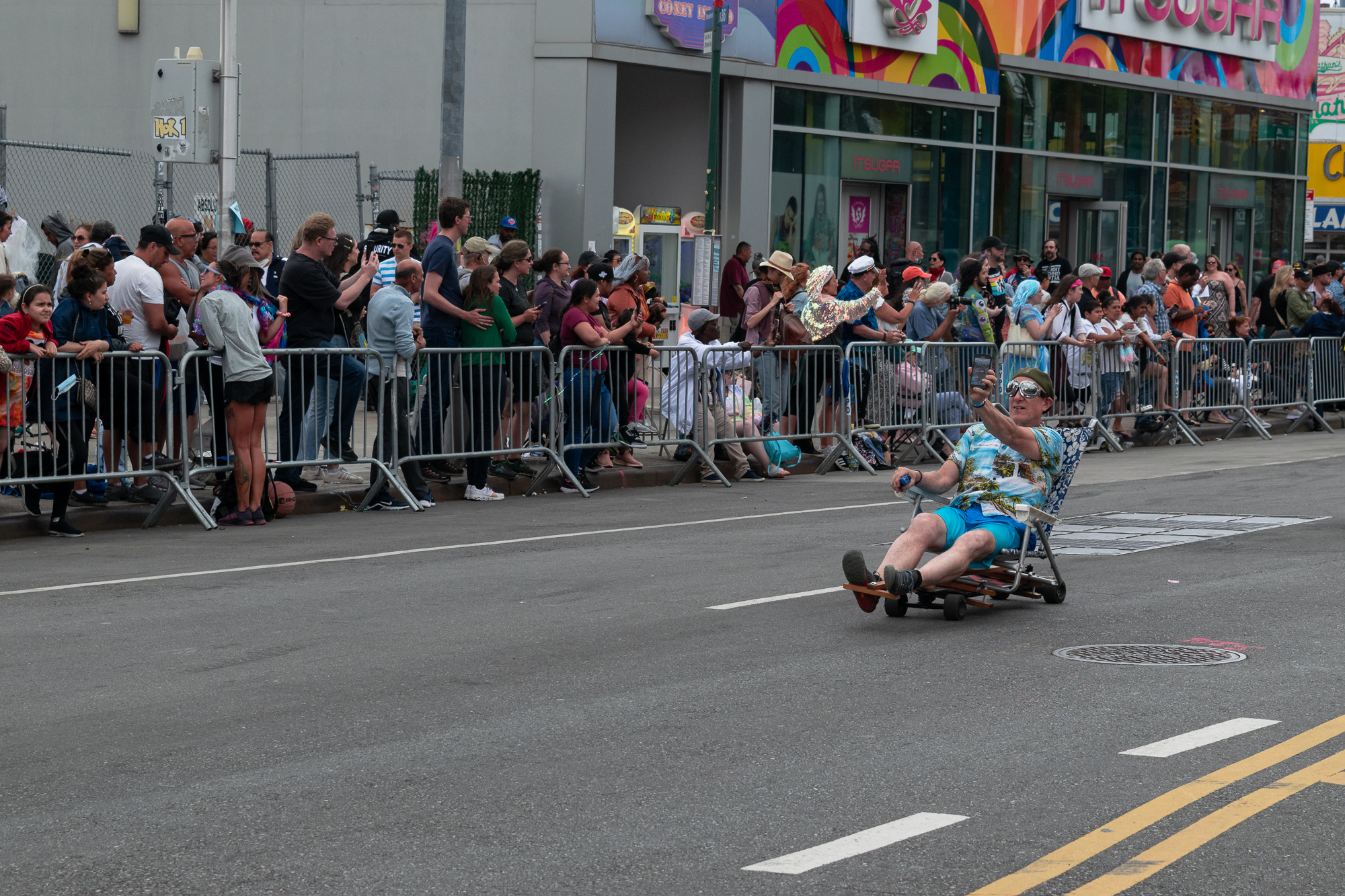 A man in a Hawaiian shirt, turquoise shorts and sunglasses reclines in a folding lawn chair mounted on a low wheeled cart, holding a phone up to film himself as he rolls down a Brooklyn street. Behind metal barricades, a large crowd watches from the sidewalk in front of the IT'SUGAR storefront.