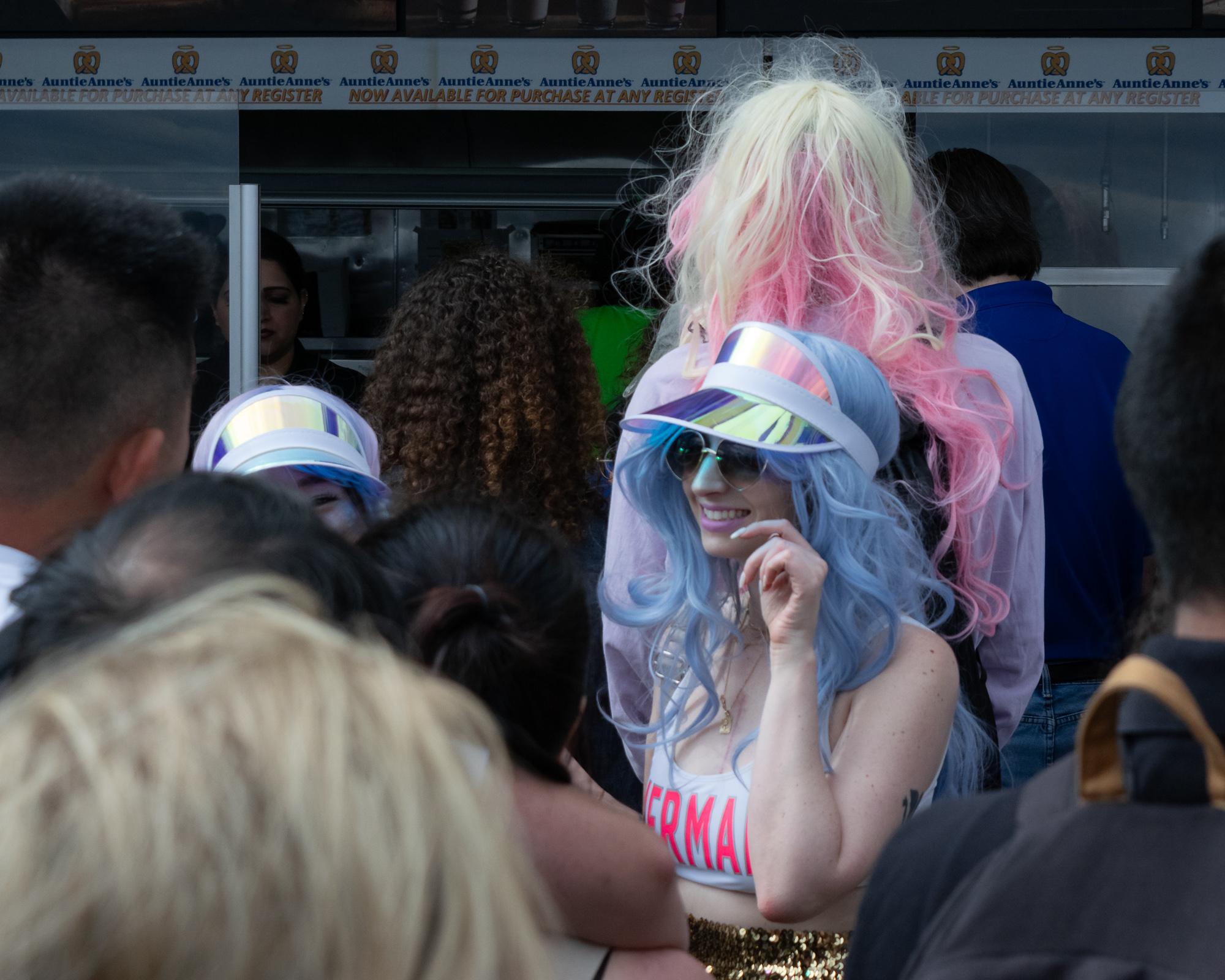 A smiling woman in a long pale blue wig, mirrored holographic visor and round sunglasses stands in a packed crowd in front of an Auntie Anne's pretzel counter, wearing a white crop top reading MERMAID and a gold sequined skirt. Behind her another person wears a towering blonde and pink wig, and a second matching visor is visible to the left.