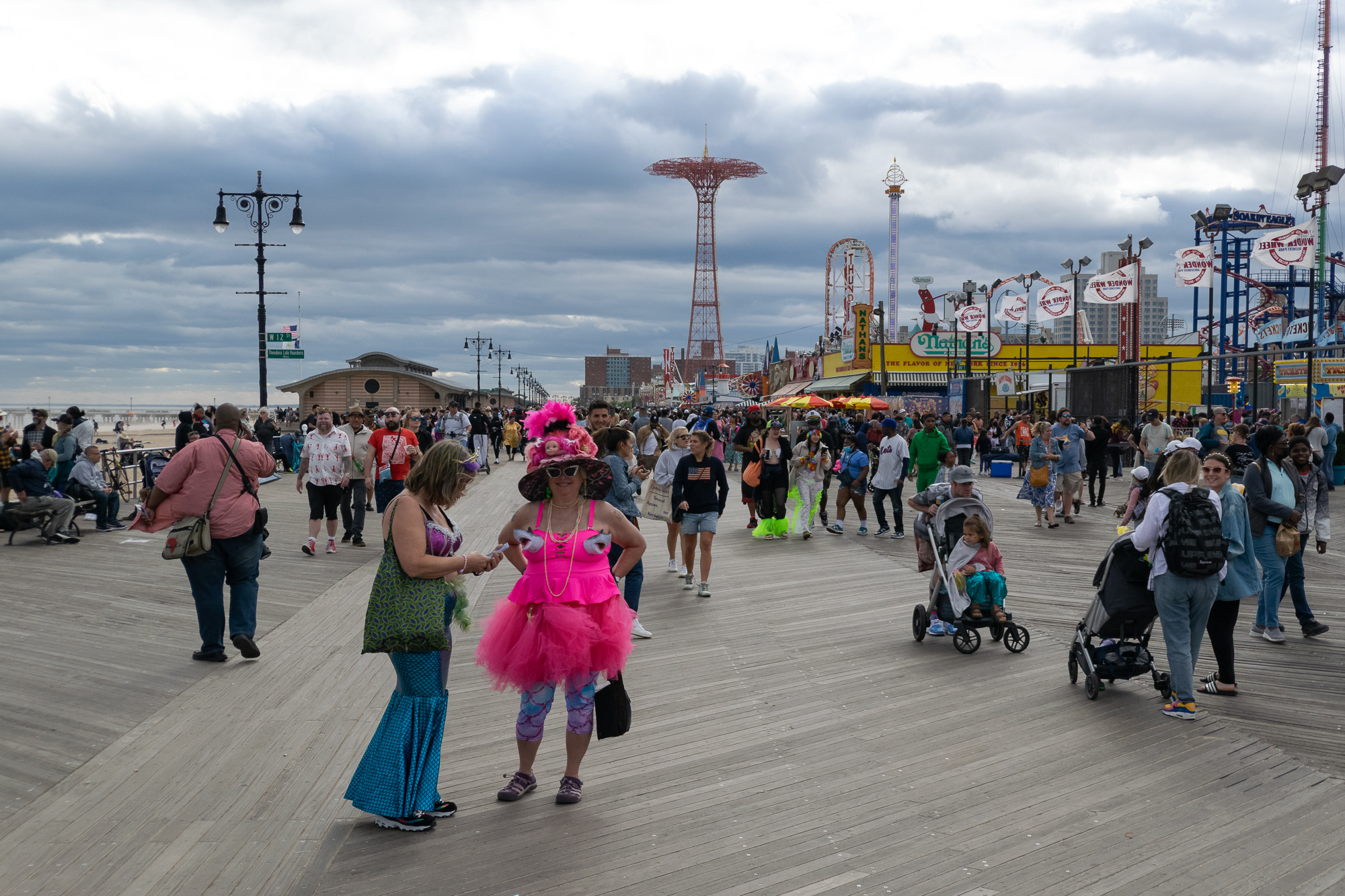 Two costumed women stand on the wide wooden boardwalk at Coney Island: one in a hot pink tutu, tank top and wide flowered hat, the other in a blue-green sequined mermaid tail skirt checking her phone. Behind them crowds walk past Nathan's Famous, the Wonder Wheel signage and the red Parachute Jump under heavy grey clouds.