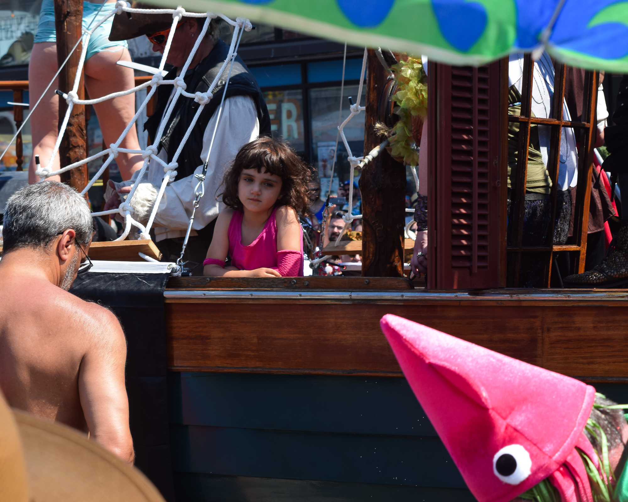 A young girl with dark curly hair in a pink tank top and pink arm warmer leans on the wooden rail of a parade float built like a pirate ship, with rope rigging and a costumed pirate behind her. In the foreground a shirtless man in sunglasses watches from below, and a bright pink fabric squid puppet with googly eyes rises at the right edge.