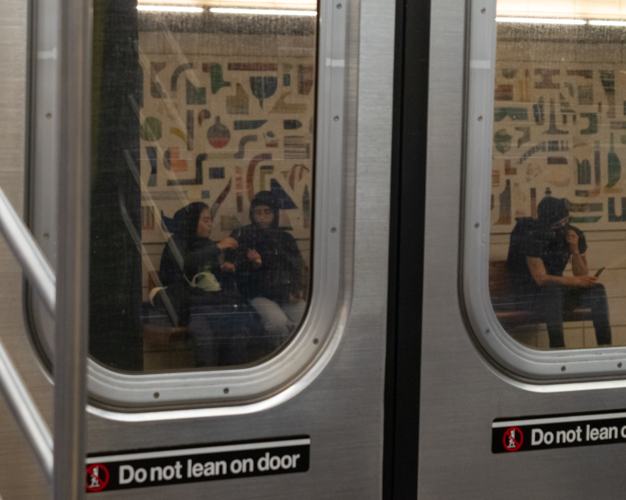 Seen from a station platform, two subway car door windows frame seated passengers inside a train: a woman in a dark jacket with a green bag beside a person in a hooded coat and glasses, and in the next window a man in a short-sleeved shirt holding a phone. A tiled mosaic wall reflects across the glass, and black stickers below the windows read Do Not Lean on Door.